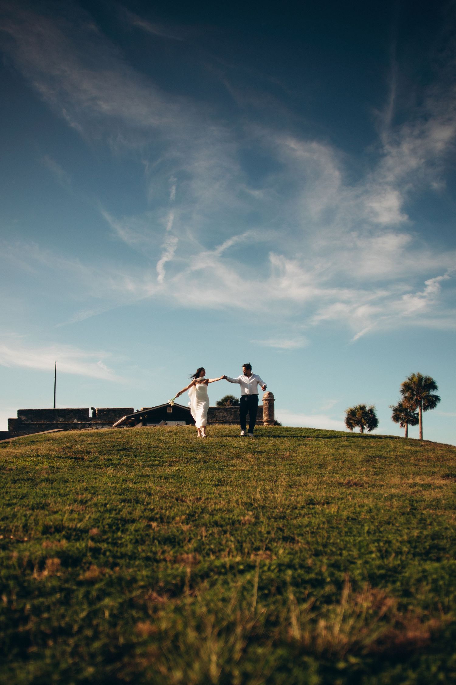 Couple holding hands on a grassy hill at sunset near historic fort with palm trees and blue sky.