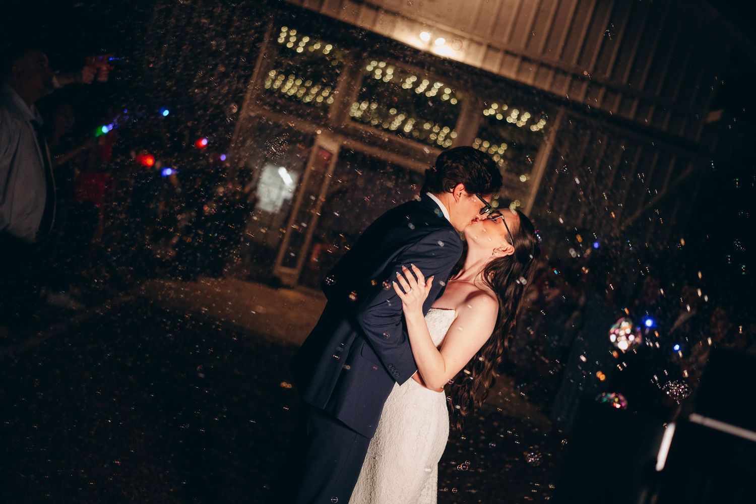 A couple shares a romantic kiss surrounded by colorful confetti outside a rustic venue at night.