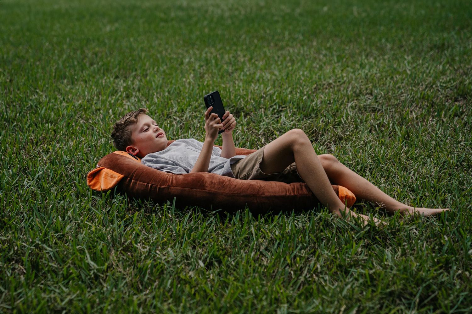 Boy lying on grass using smartphone, resting on orange pillow outdoors in green lawn.