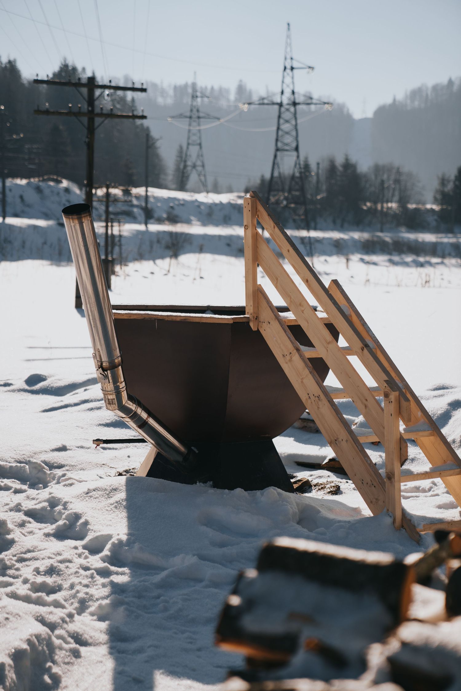 Rustic outdoor wooden hot tub with stairs in snowy winter landscape near power lines.