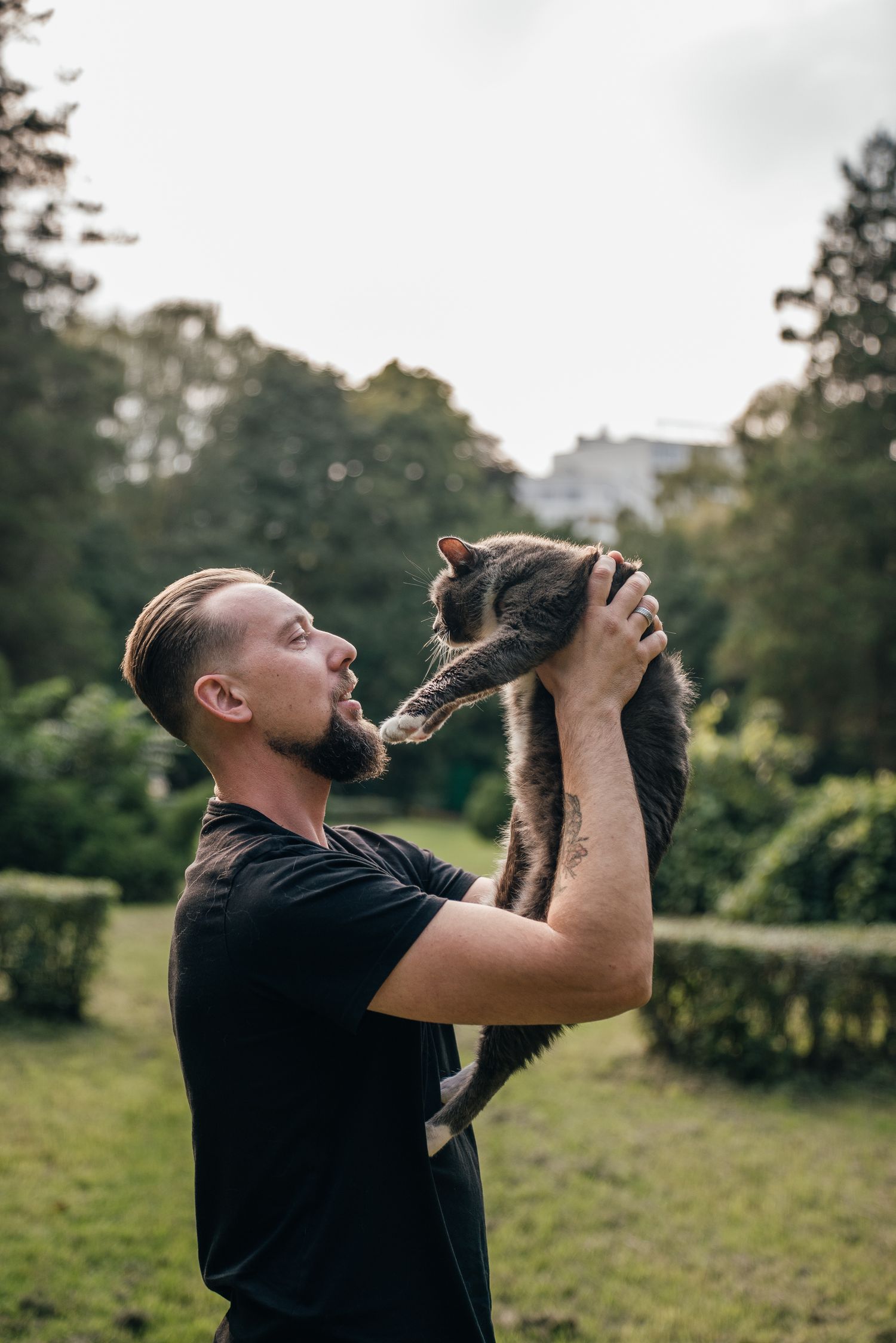 Man in black t-shirt holds gray cat above his head in a lush green park.