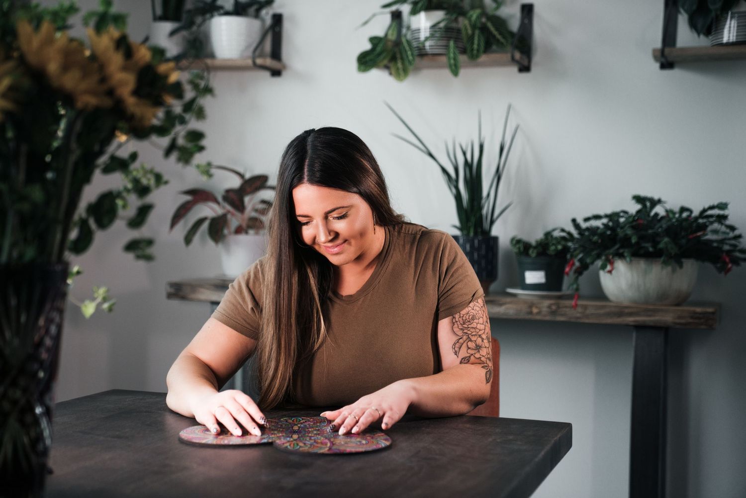 Woman with tattoos smiling while assembling a circular wooden puzzle at a table surrounded by green plants.