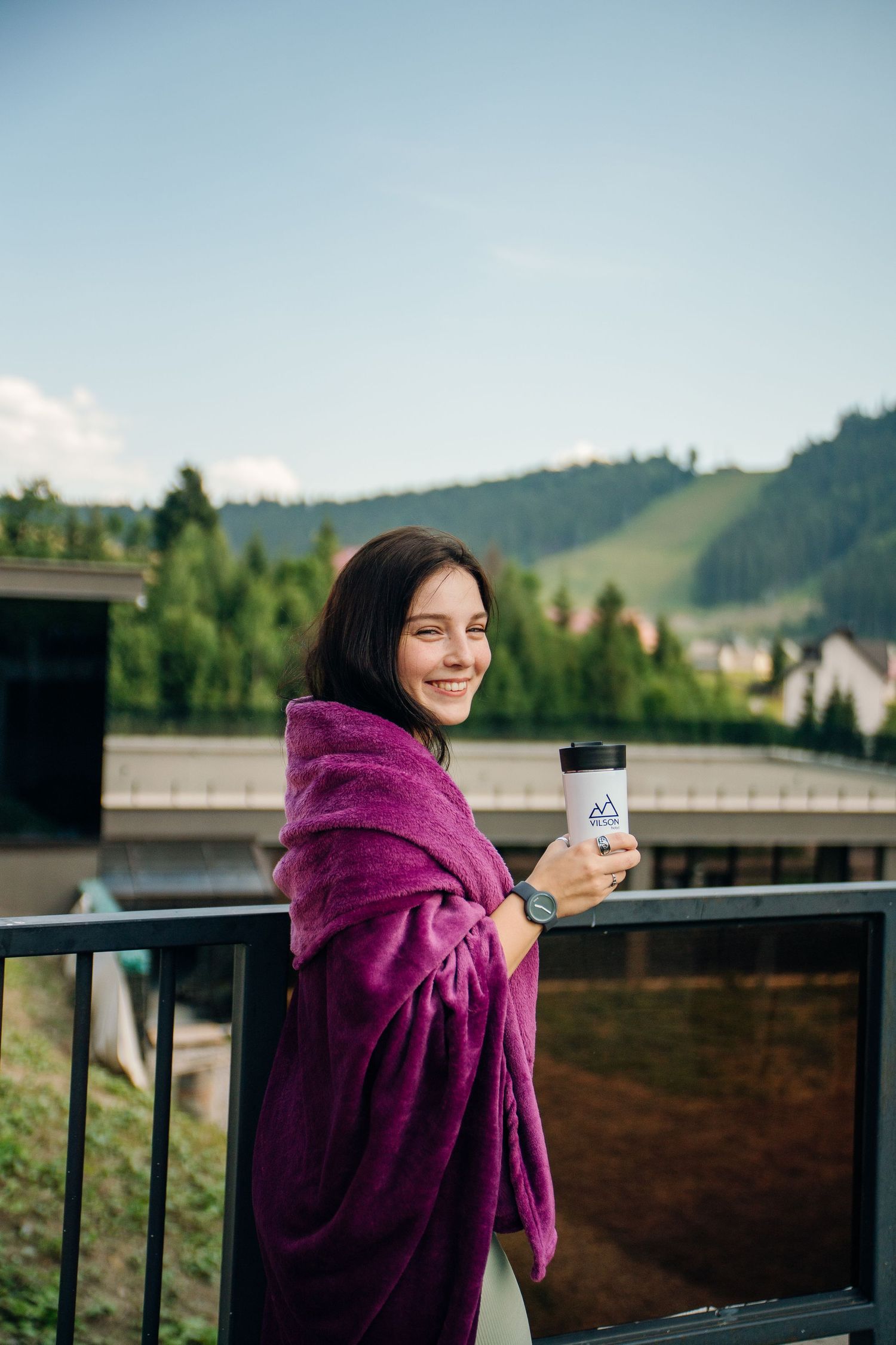 Smiling woman wrapped in purple blanket holding a travel cup on a balcony with mountain forest views.