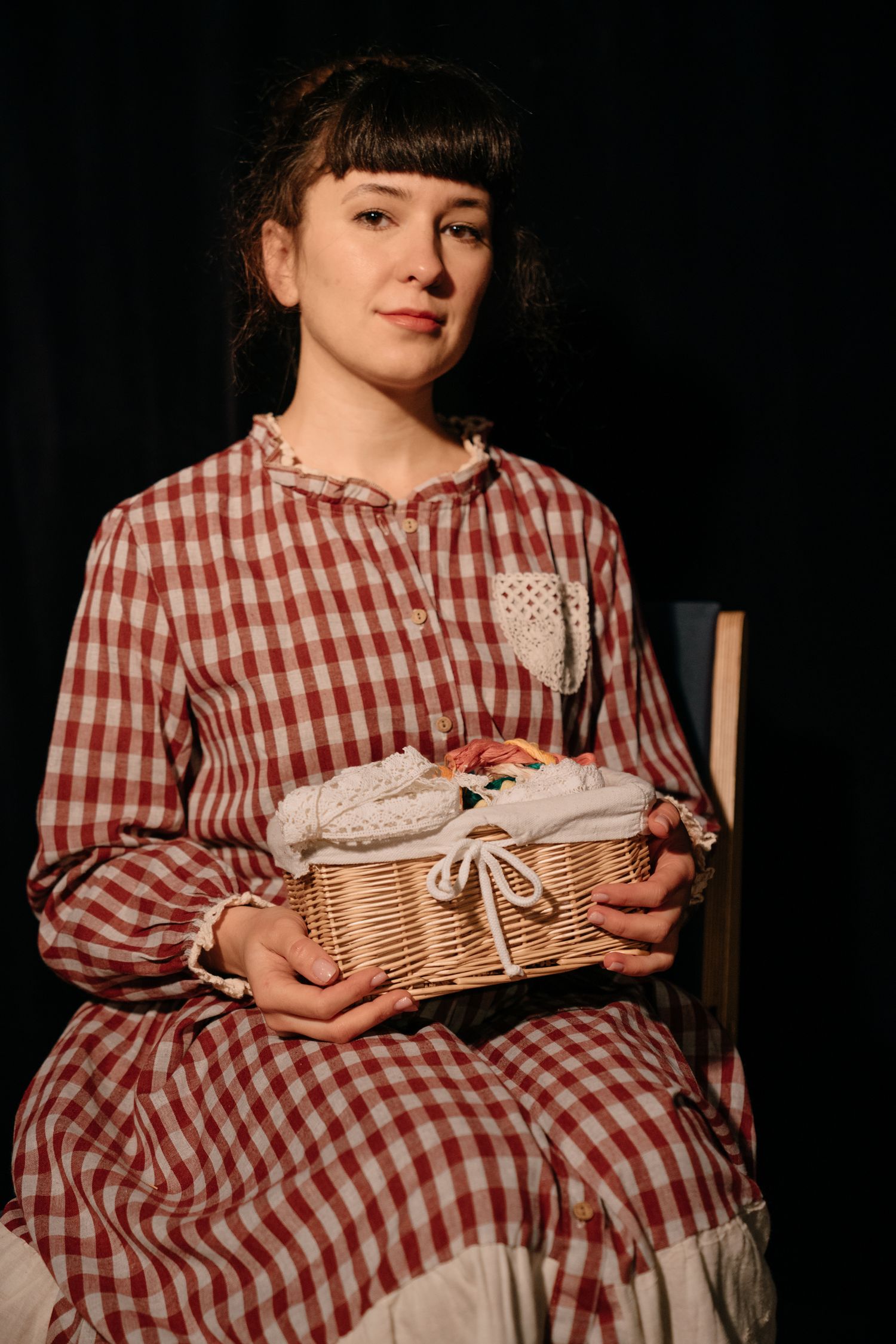Woman in vintage red gingham dress holding a wicker basket while seated against dark background.