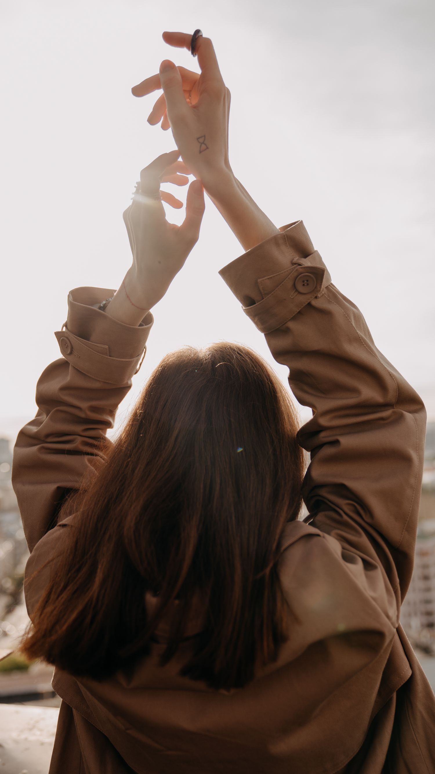 Woman in beige trench coat raising hands toward bright sky, back view.