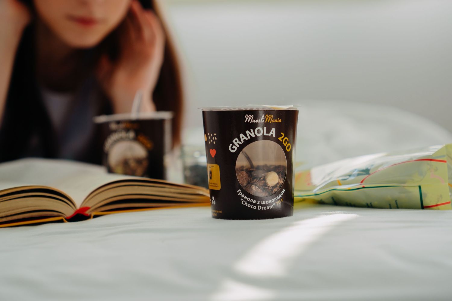 Cup of granola on a bed next to an open book with a blurred person reading in background.