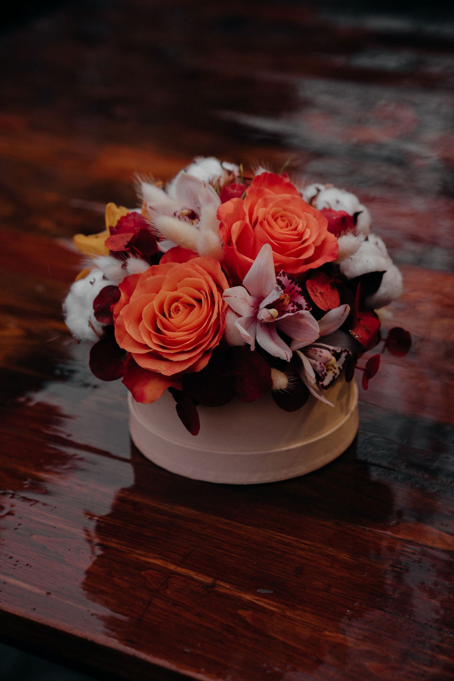 Elegant floral arrangement with orange roses, cotton flowers, and orchids in a white round box on a wooden surface.