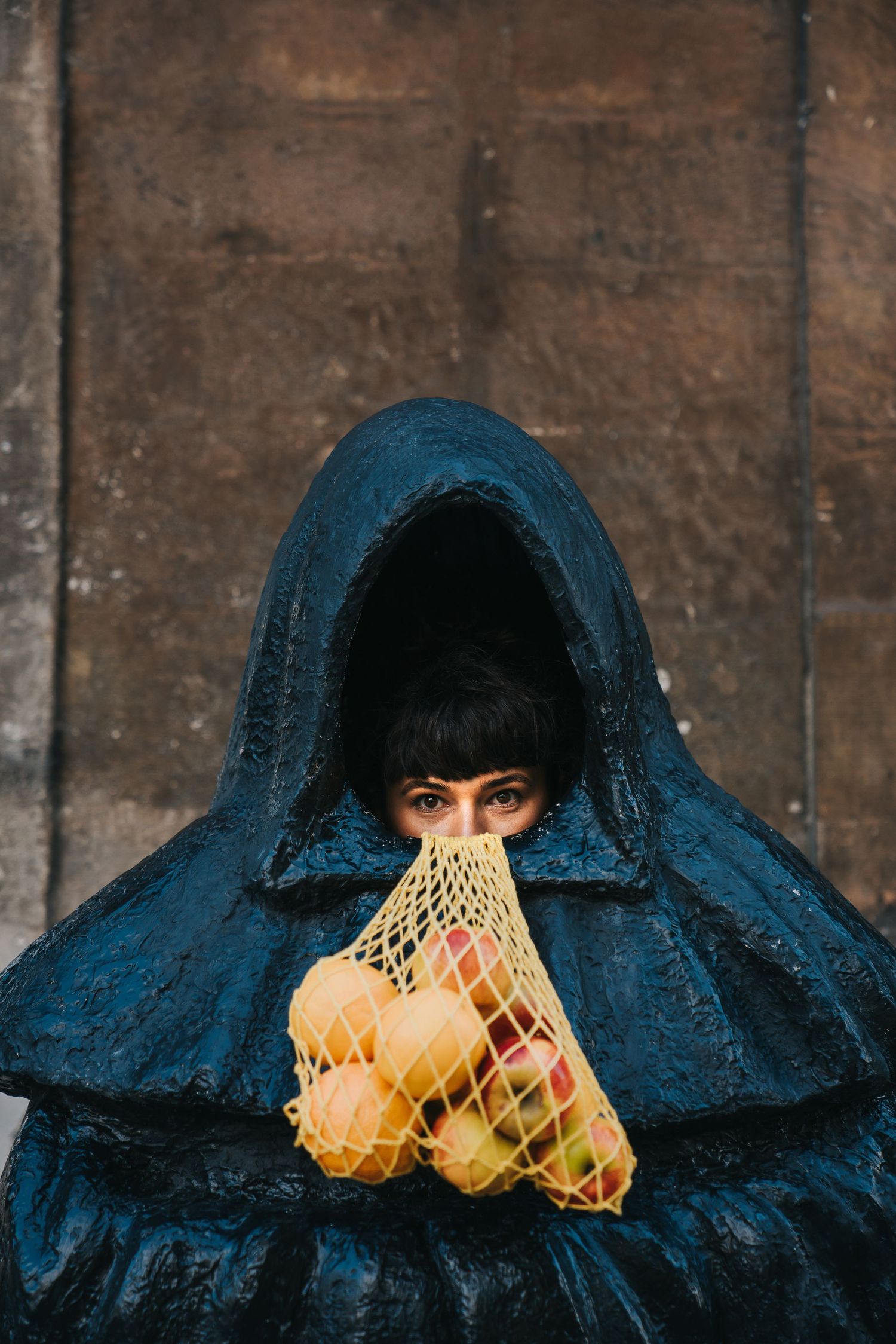 Person in dark hooded cloak holding mesh bag with fresh fruits near stone wall.