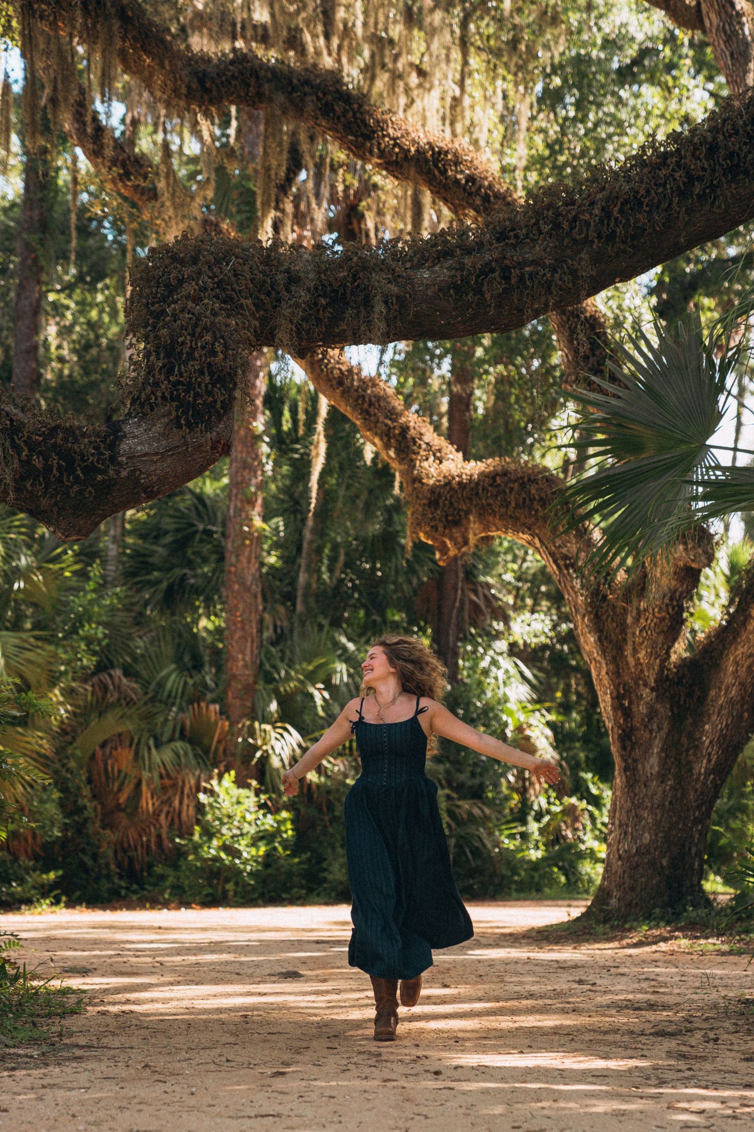 A person in a dark dress dances along a scenic outdoor path surrounded by moss-draped oak trees.