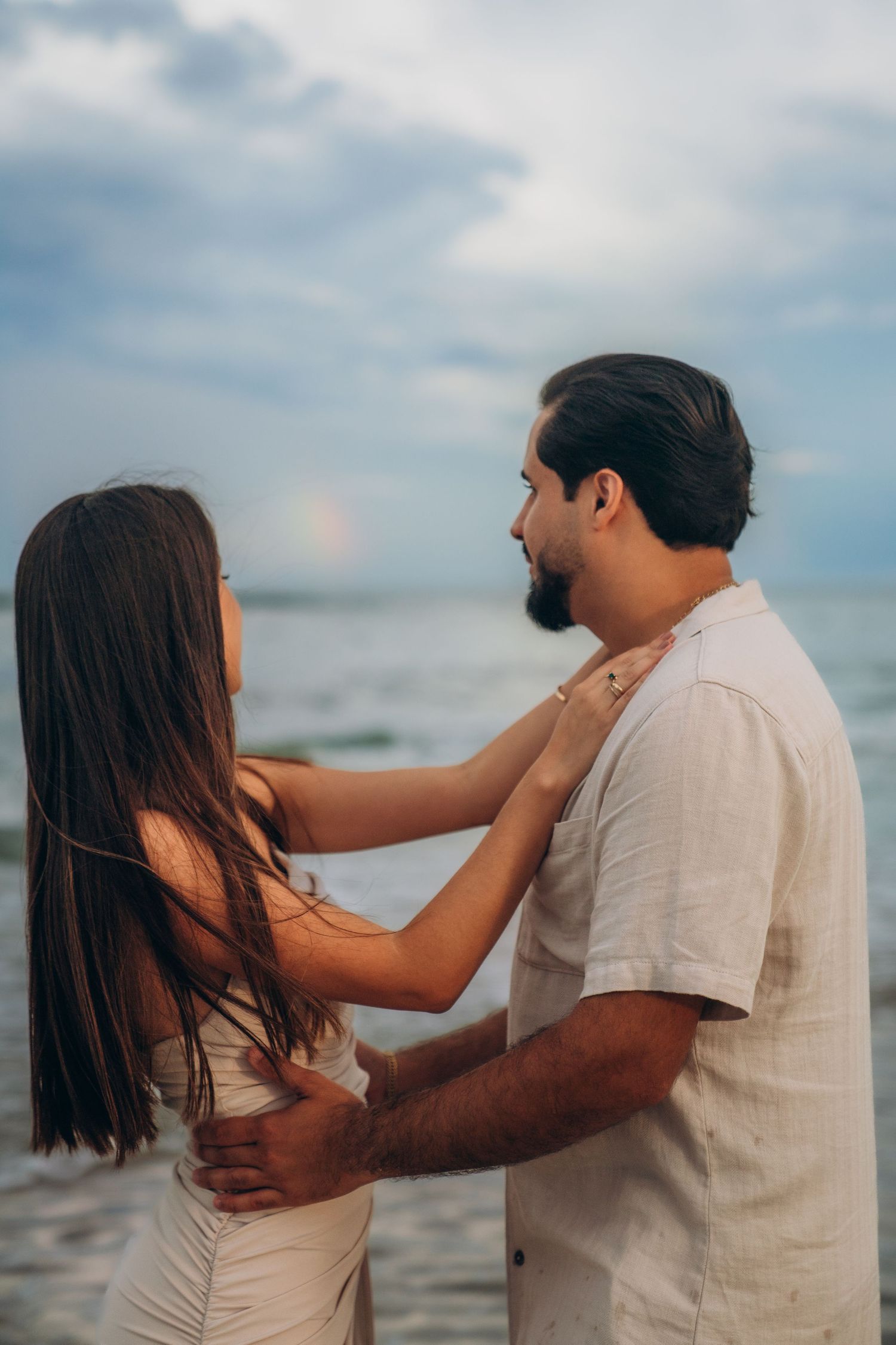 Playful couple in white outfits sharing intimate moments on a beach at sunset.