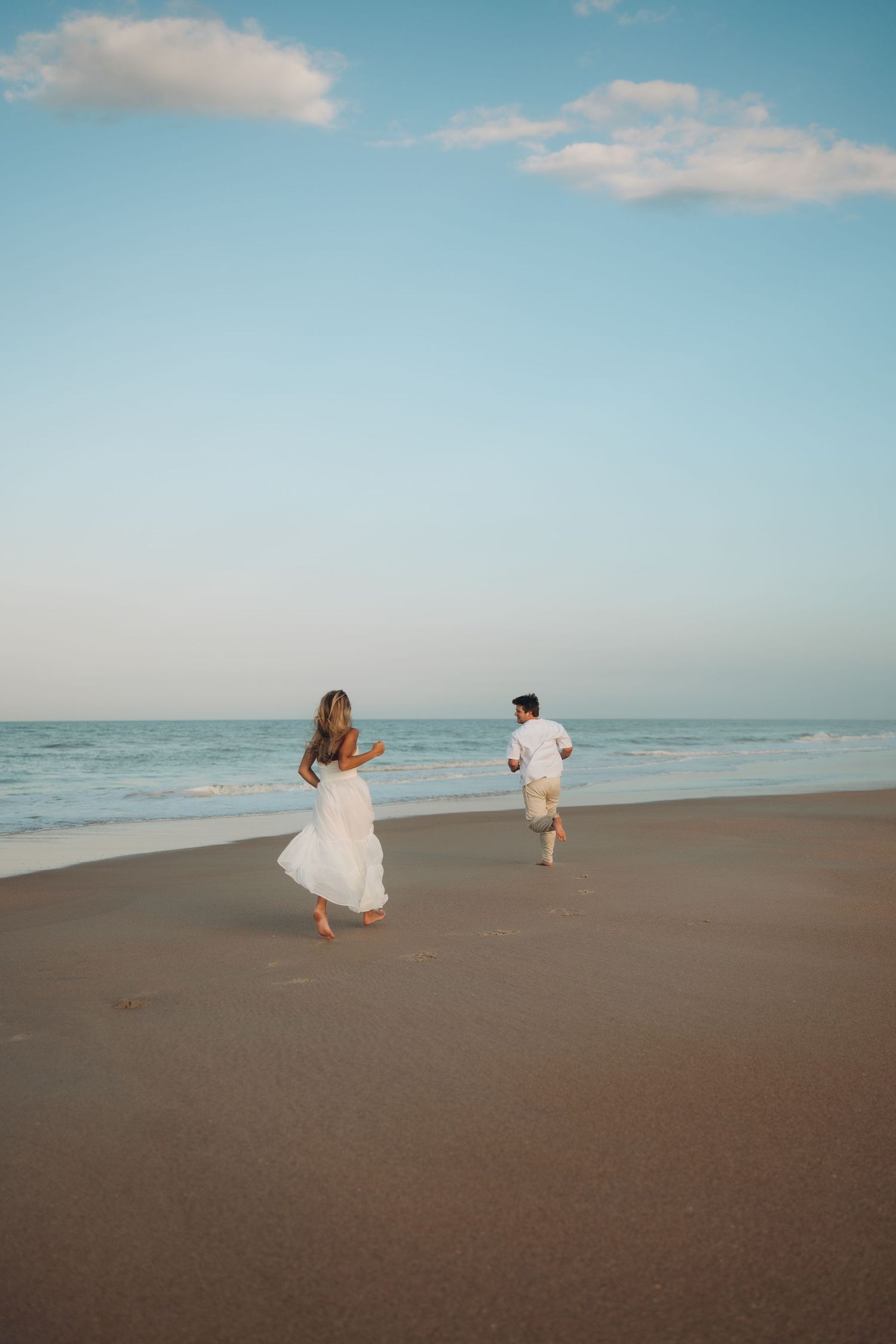 A couple runs playfully along an empty beach with blue skies overhead.