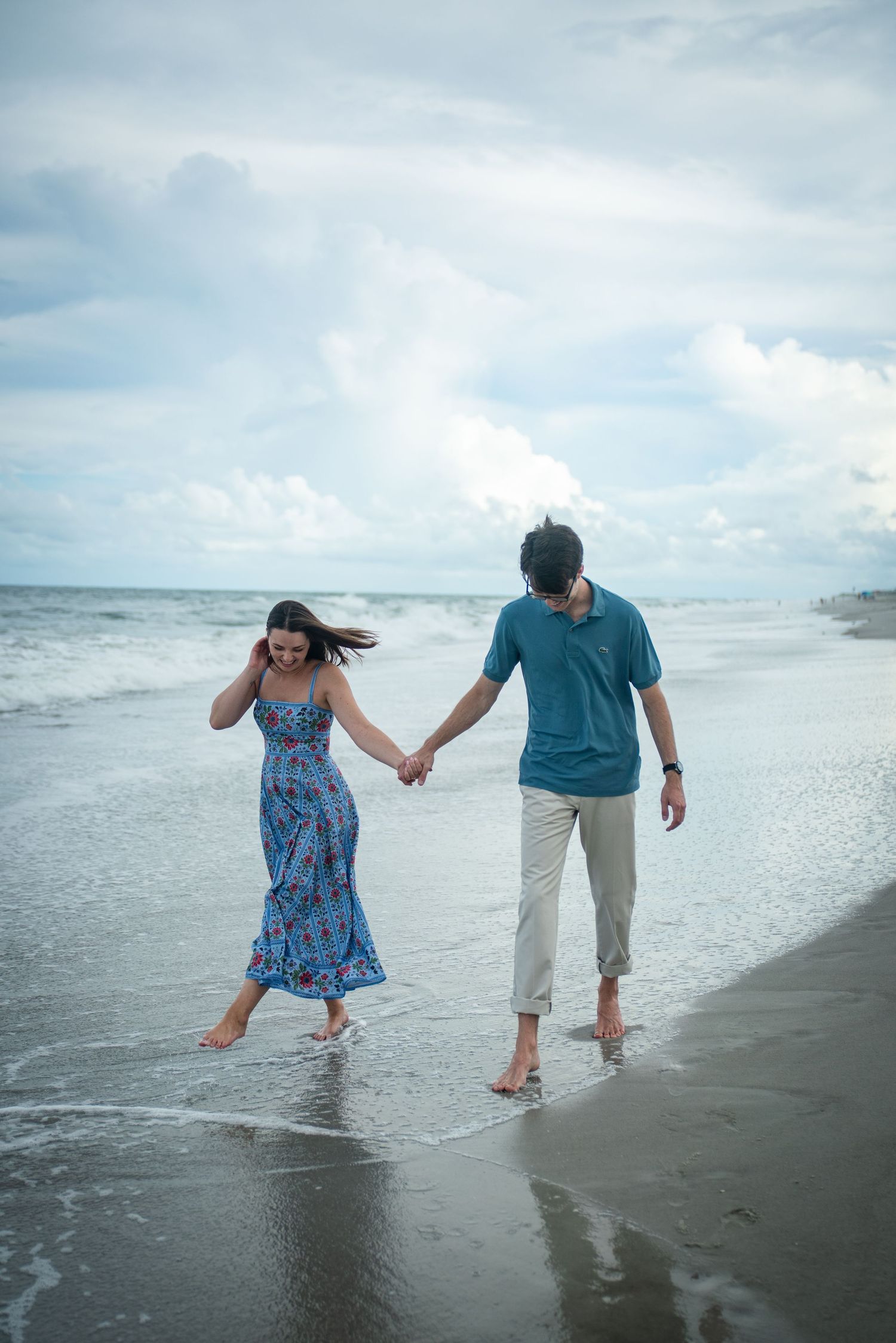 A couple walking hand in hand along the beach shore with waves crashing beside them.