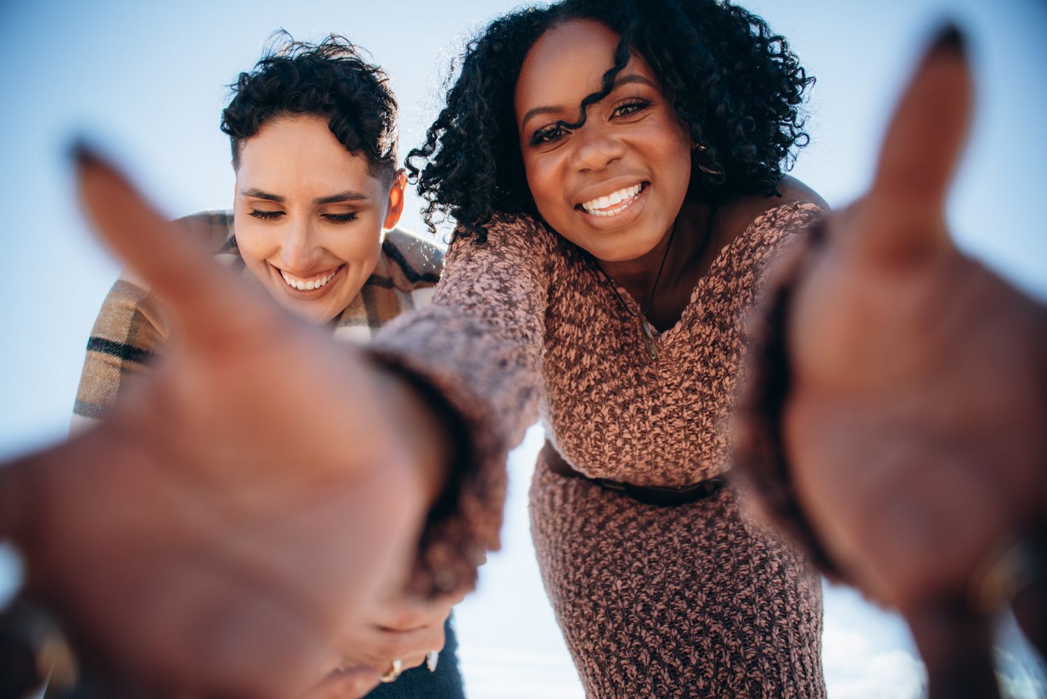 Two smiling women pointing fingers at camera against blue sky background.
