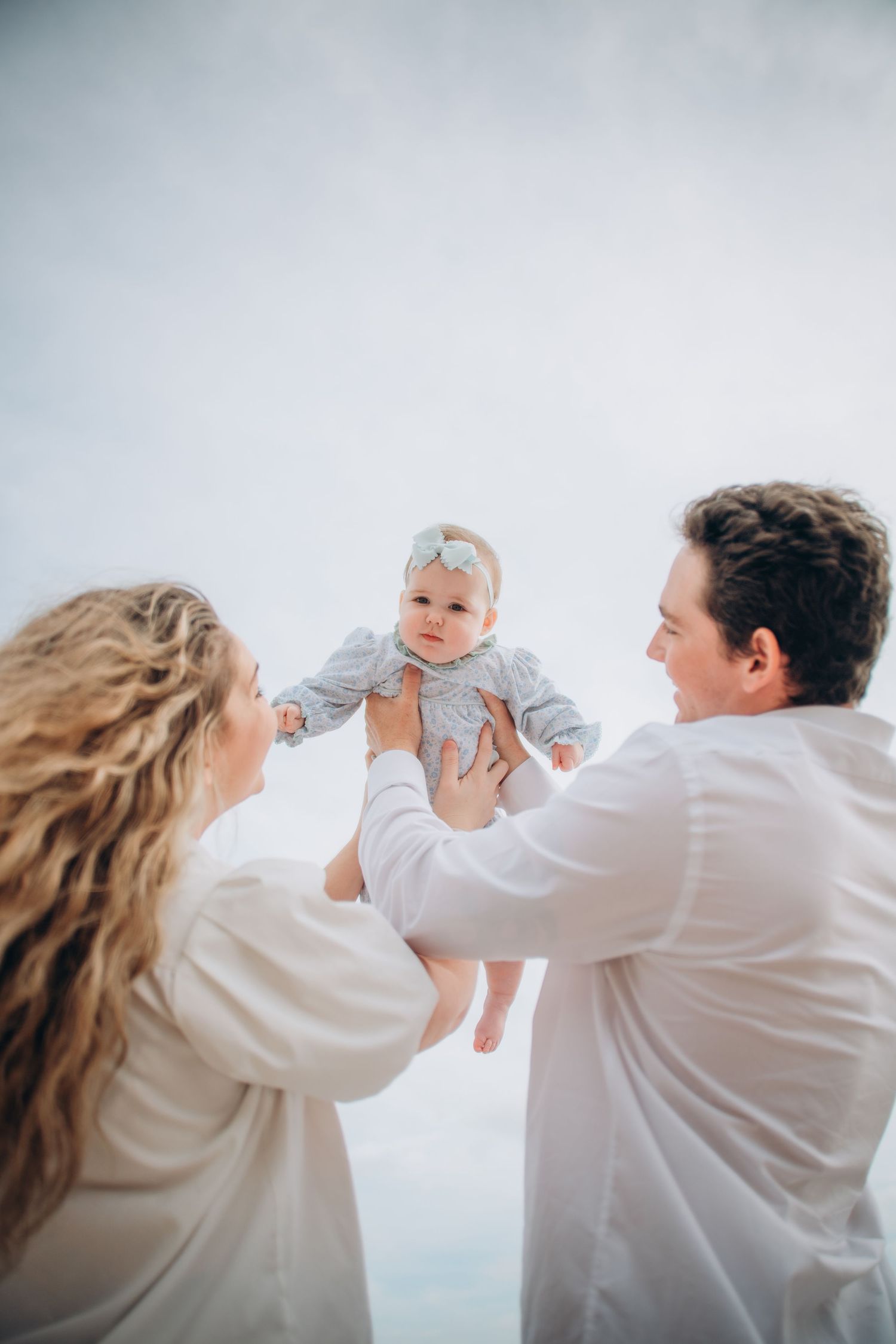 Happy parents in white shirts lifting their baby girl outdoors against a bright sky.