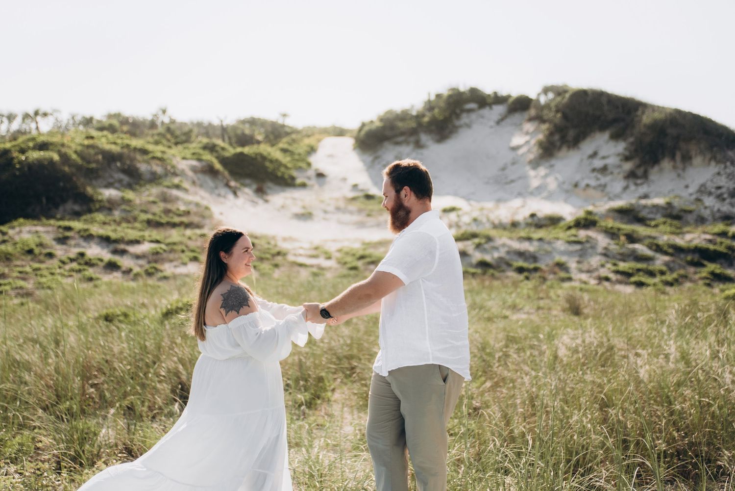Couple in white outfits holding hands in sandy dunes with grass.