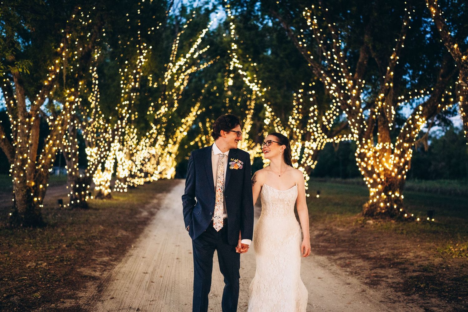 Bride and groom walk hand-in-hand down a romantic tree-lined path adorned with twinkling fairy lights at dusk.