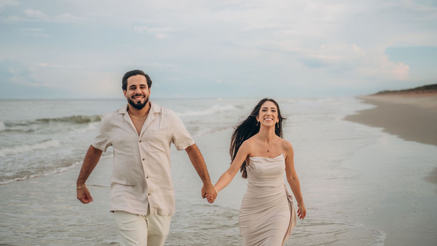 Couple walking hand in hand along a scenic beach shore at sunset wearing light colored outfits.