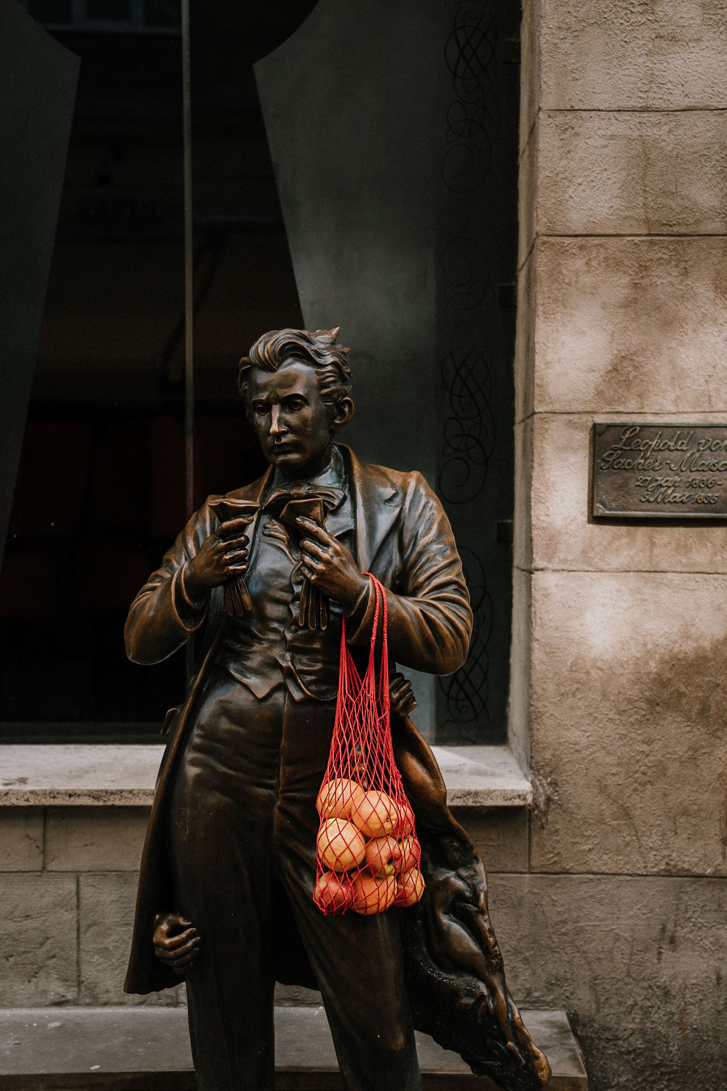 Bronze statue of a man holding a red mesh bag filled with apples beside a stone building.