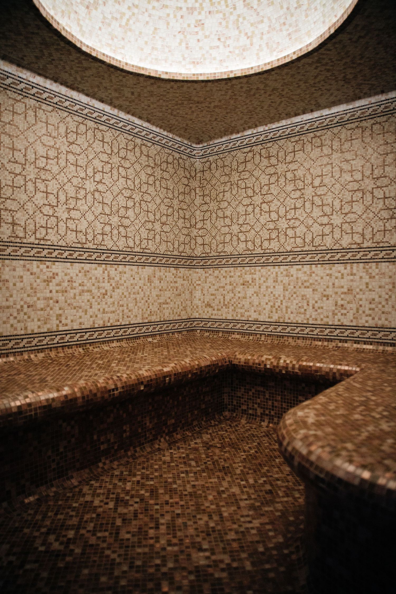 Elegant Turkish hammam steam room with mosaic tile walls, built-in marble benches, and domed ceiling with circular skylight.