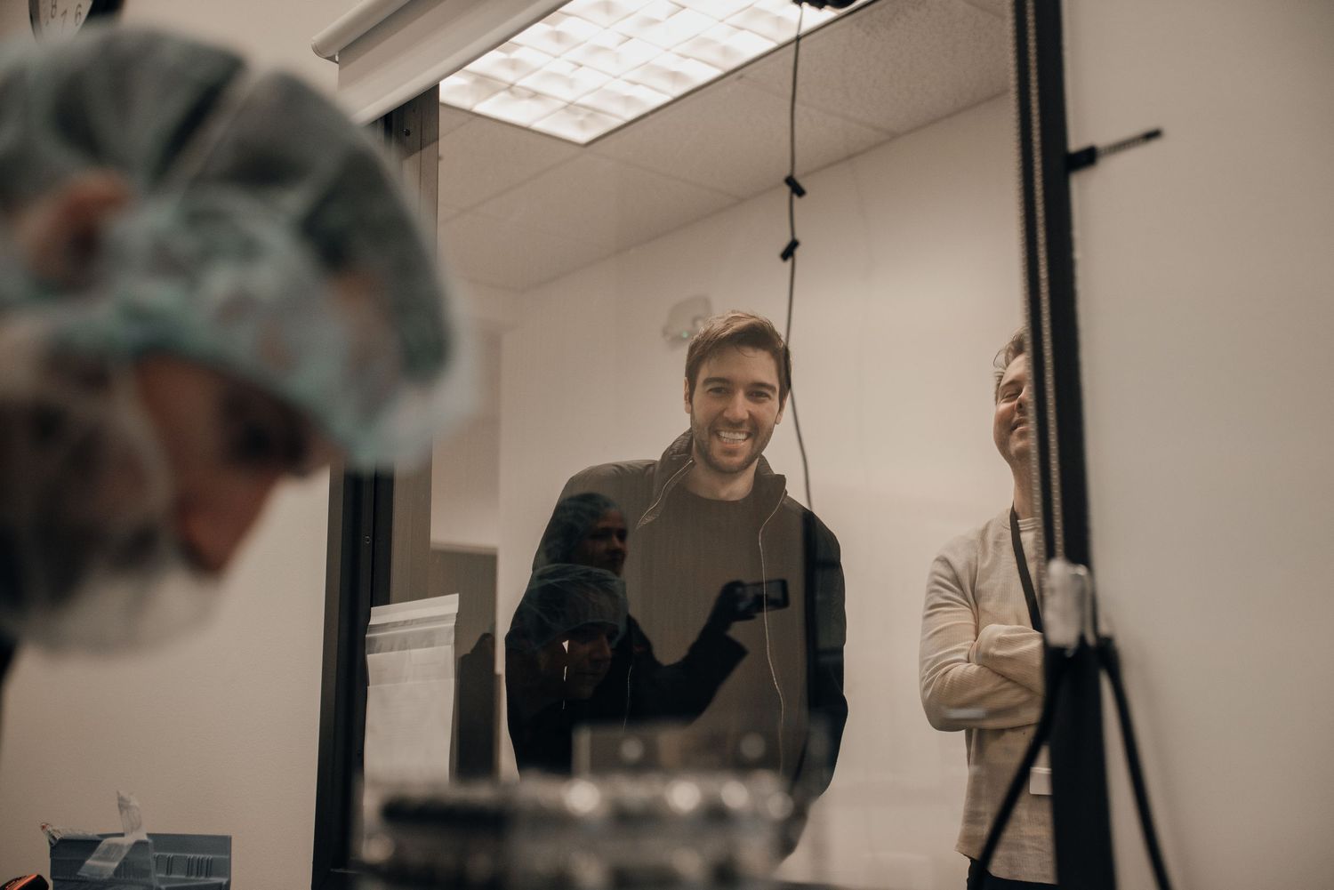 Smiling man in a clean room facility wearing protective gear, standing near a glass partition with colleagues.