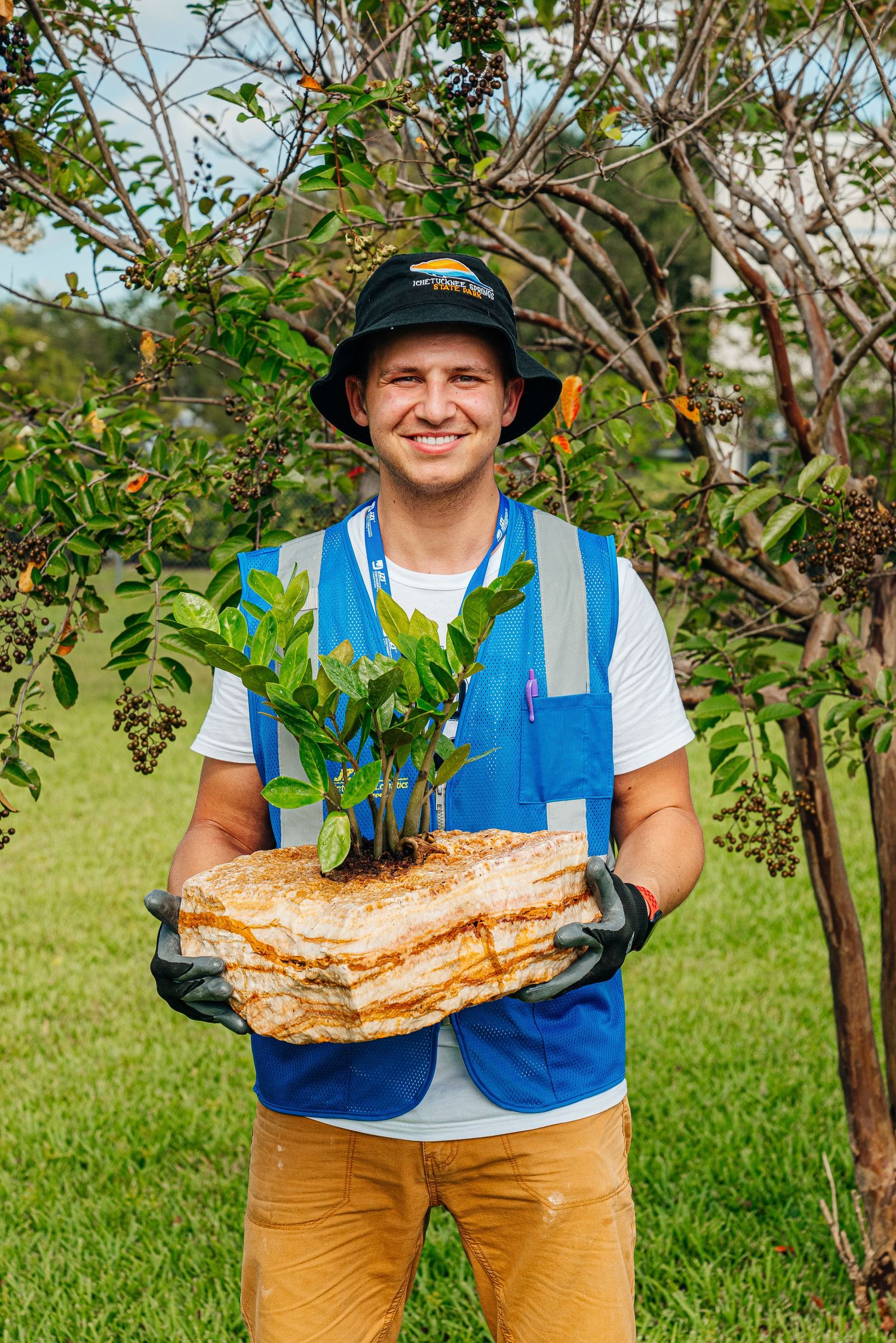 Smiling worker in blue vest holding plant on layered rock in garden setting.