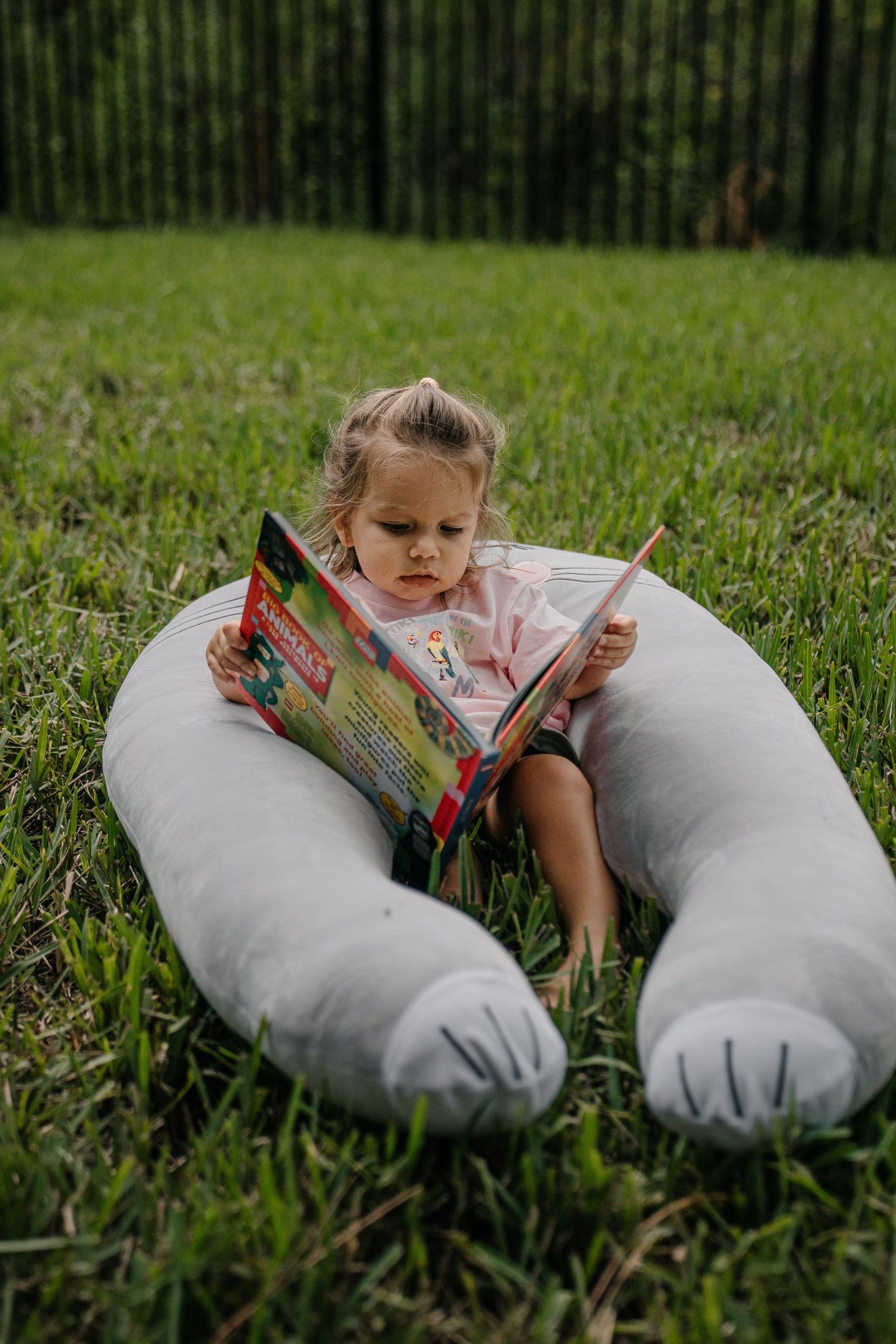 Young girl reading a colorful book outdoors, sitting in a large U-shaped gray pillow on green grass.