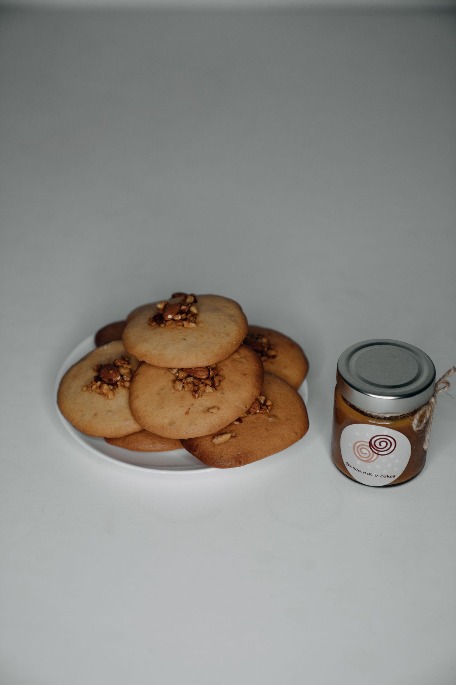 Plate of walnut-topped butter cookies beside a small jar of nut spread on a white surface.