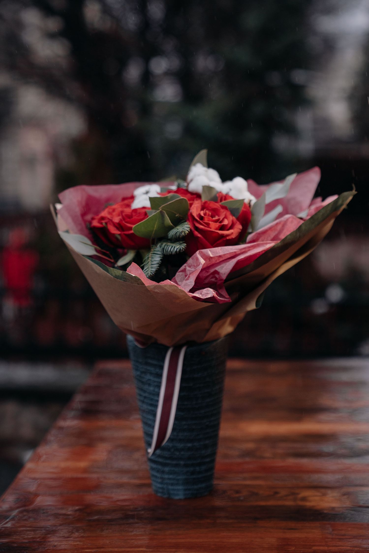 Elegant red roses bouquet wrapped in pink paper, displayed in a vase on a wooden table with bokeh background.