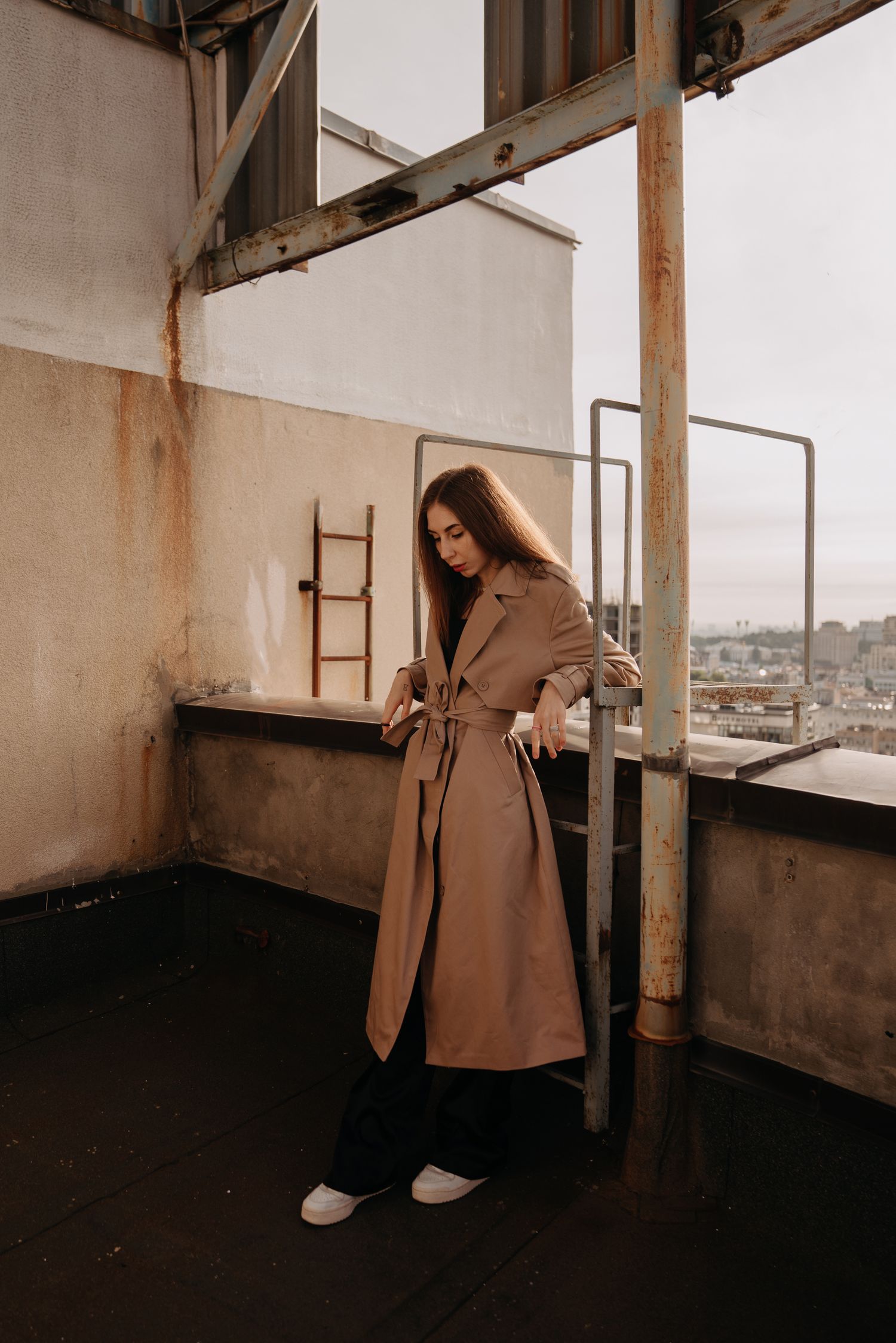 Woman in beige trench coat posing on urban rooftop with city skyline backdrop.
