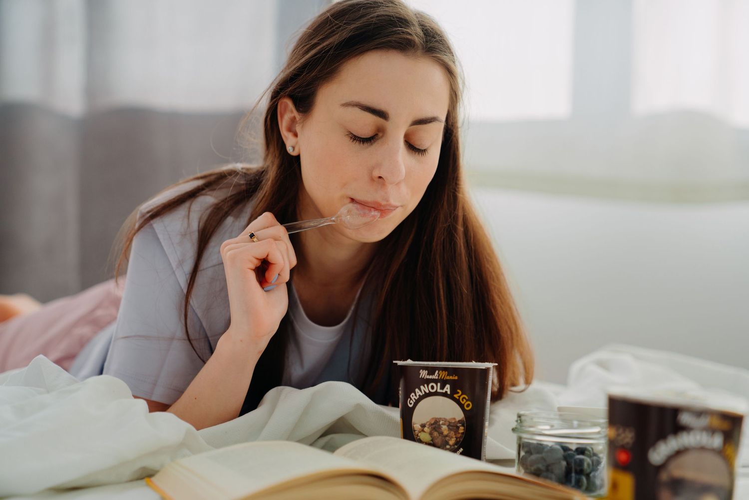Young woman eating granola in bed while reading a book, enjoying a relaxing morning snack.
