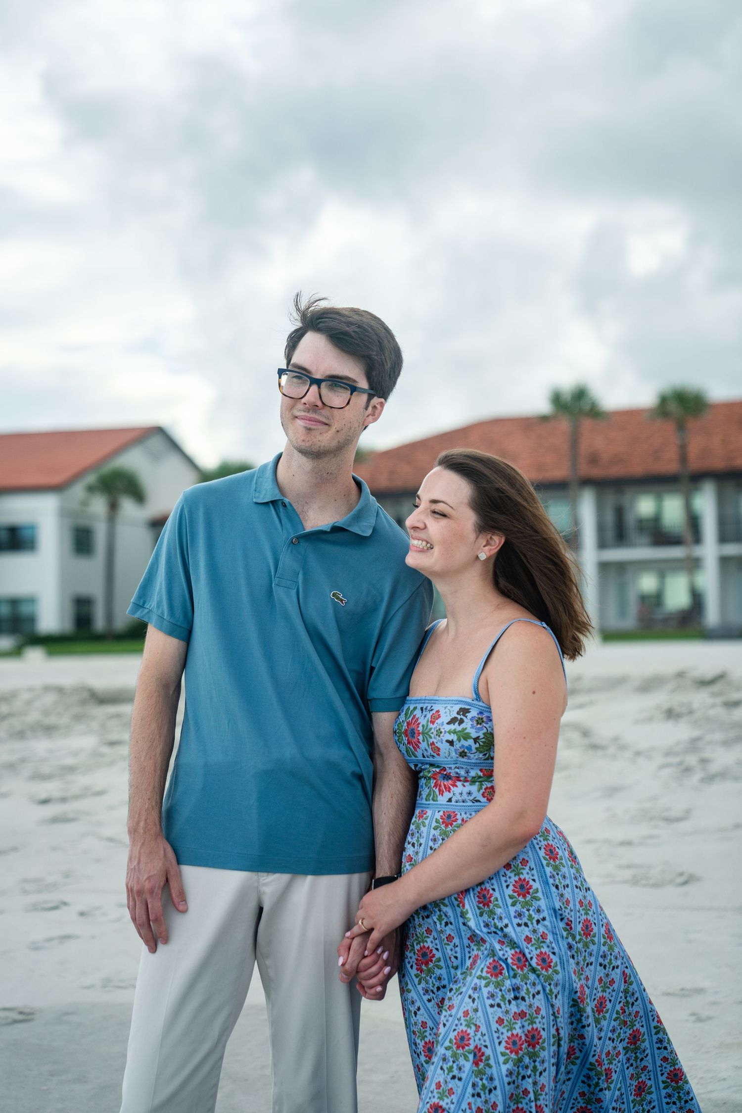 A couple poses together on the beach with coastal buildings visible in the background.