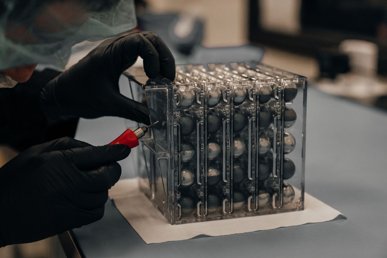 Technician in black gloves assembling a cylindrical battery cell module with a red tool in a lab.