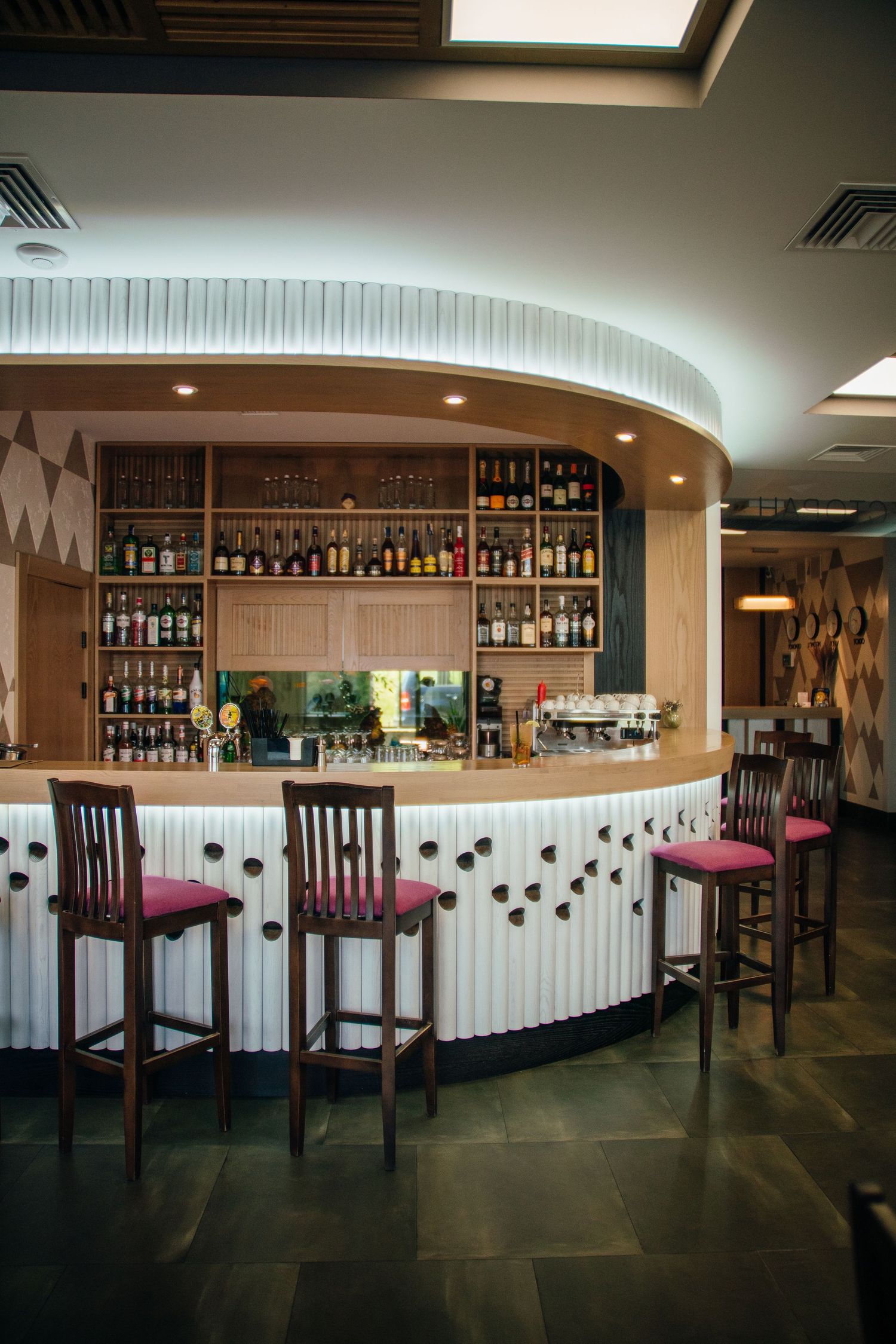 Elegant curved bar with wood accents, pink bar stools, and backlit shelves stocked with bottles in a modern lounge.