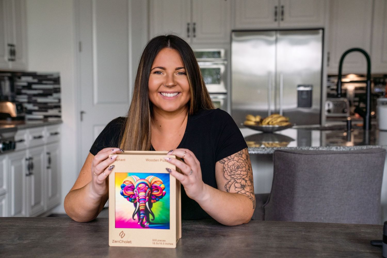 Smiling woman with tattoos holds colorful elephant artwork in a modern white kitchen.