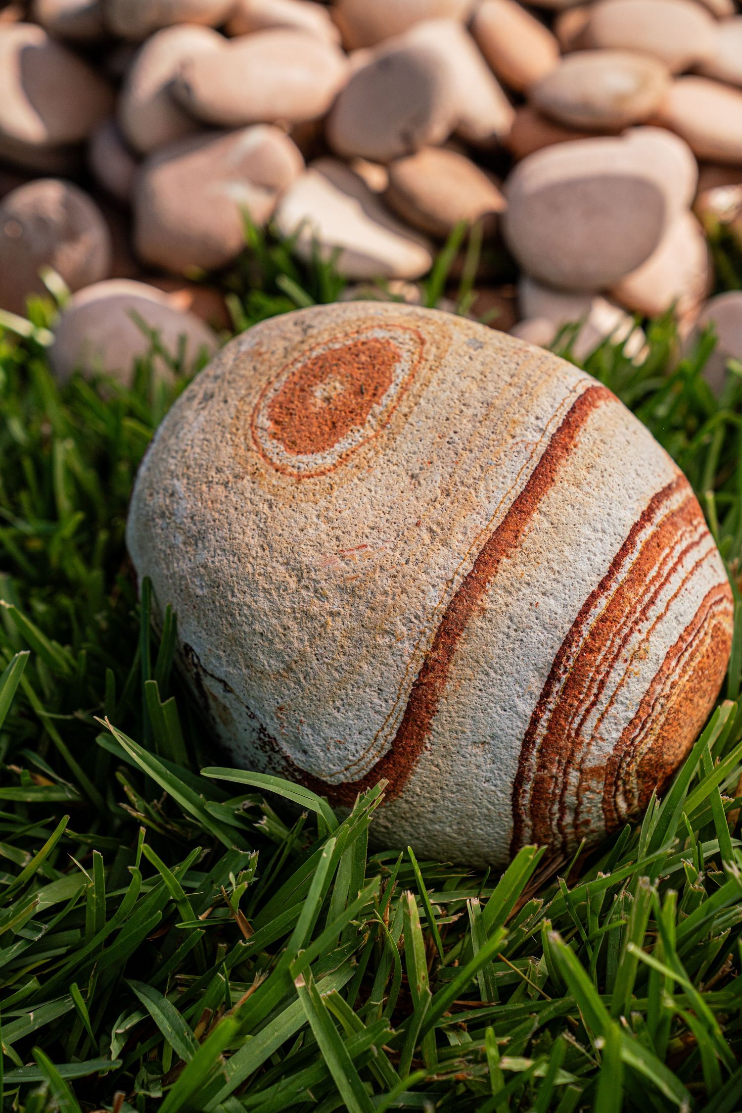 Large oval rock with red and orange circular stripe patterns sitting on green grass with pebbles.