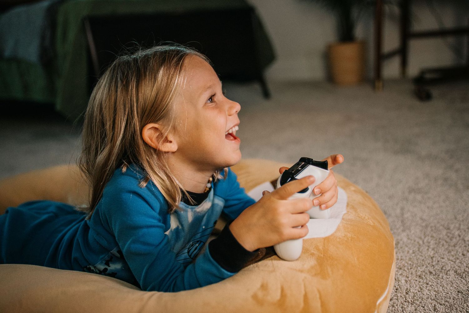 Young child lying on a bean bag chair playing video games with a controller, smiling happily.