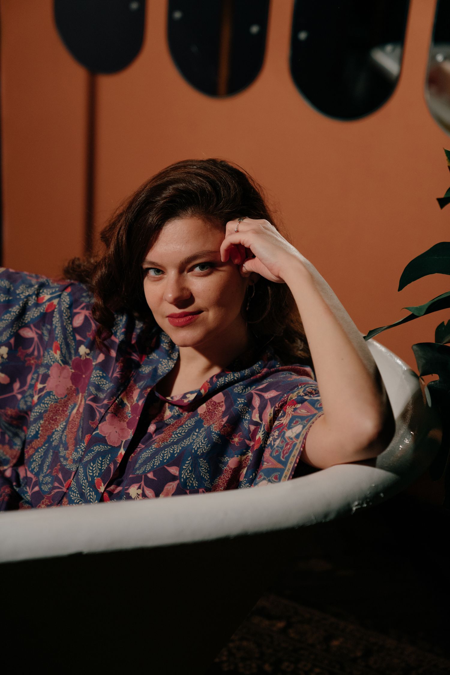 Woman in floral shirt relaxing in a vintage bathtub, smiling softly against a warm terracotta background.