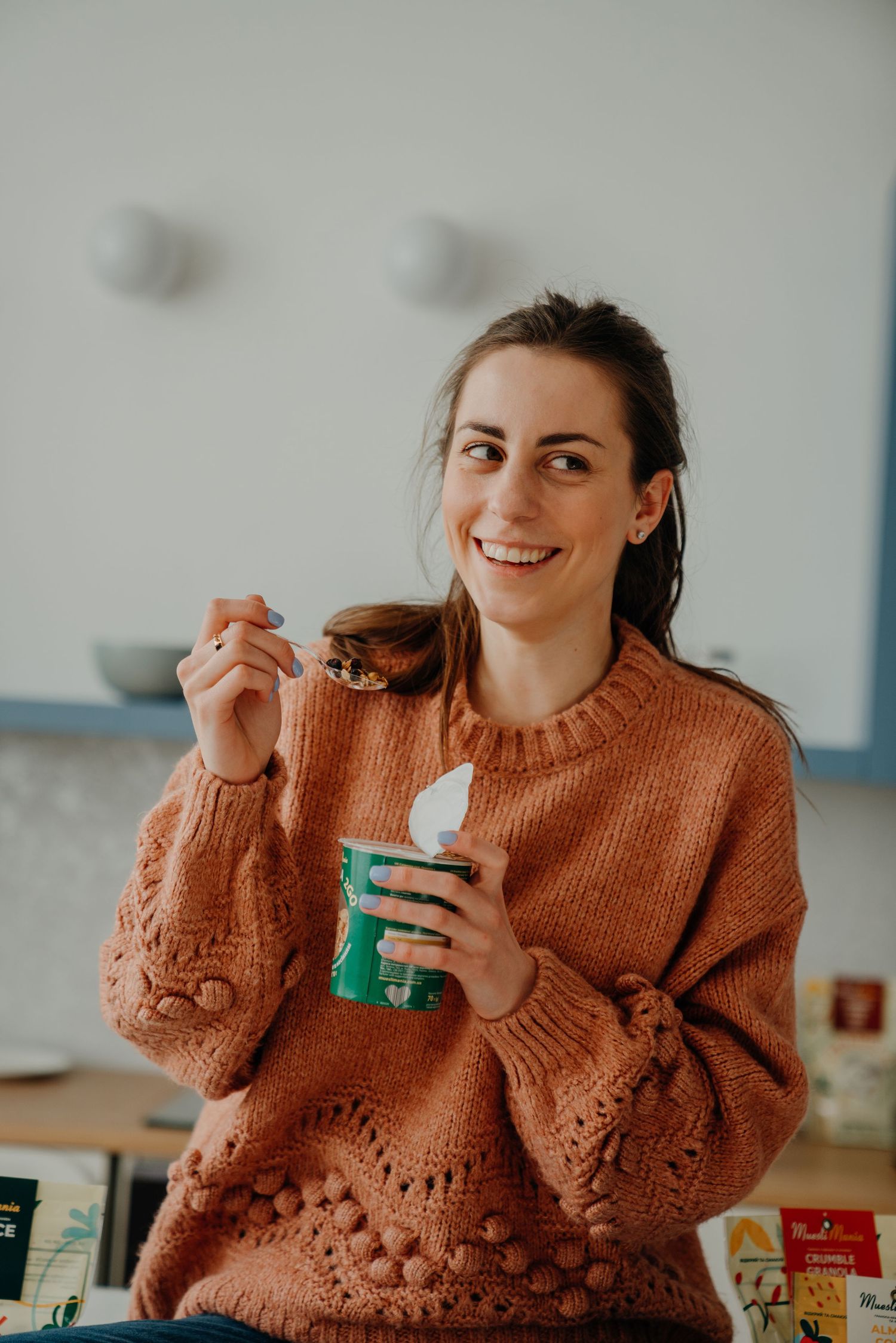 A smiling woman in an orange knit sweater enjoying a cup of granola in a bright kitchen.