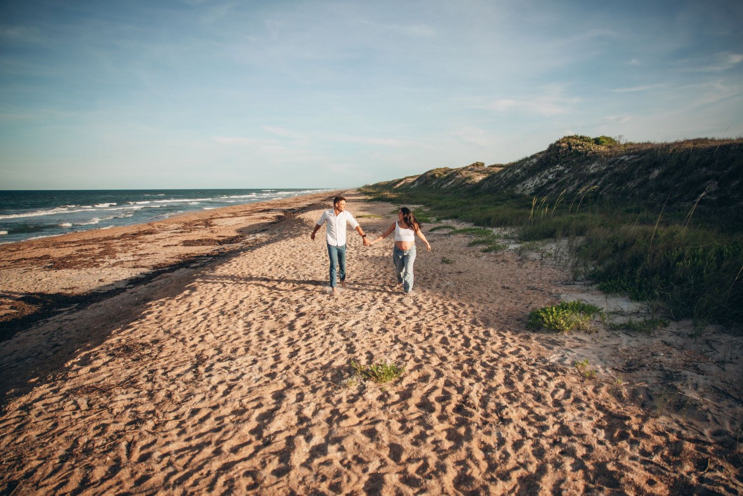 Couple holding hands walking on a sandy beach with ocean waves and green dunes under a blue sky.
