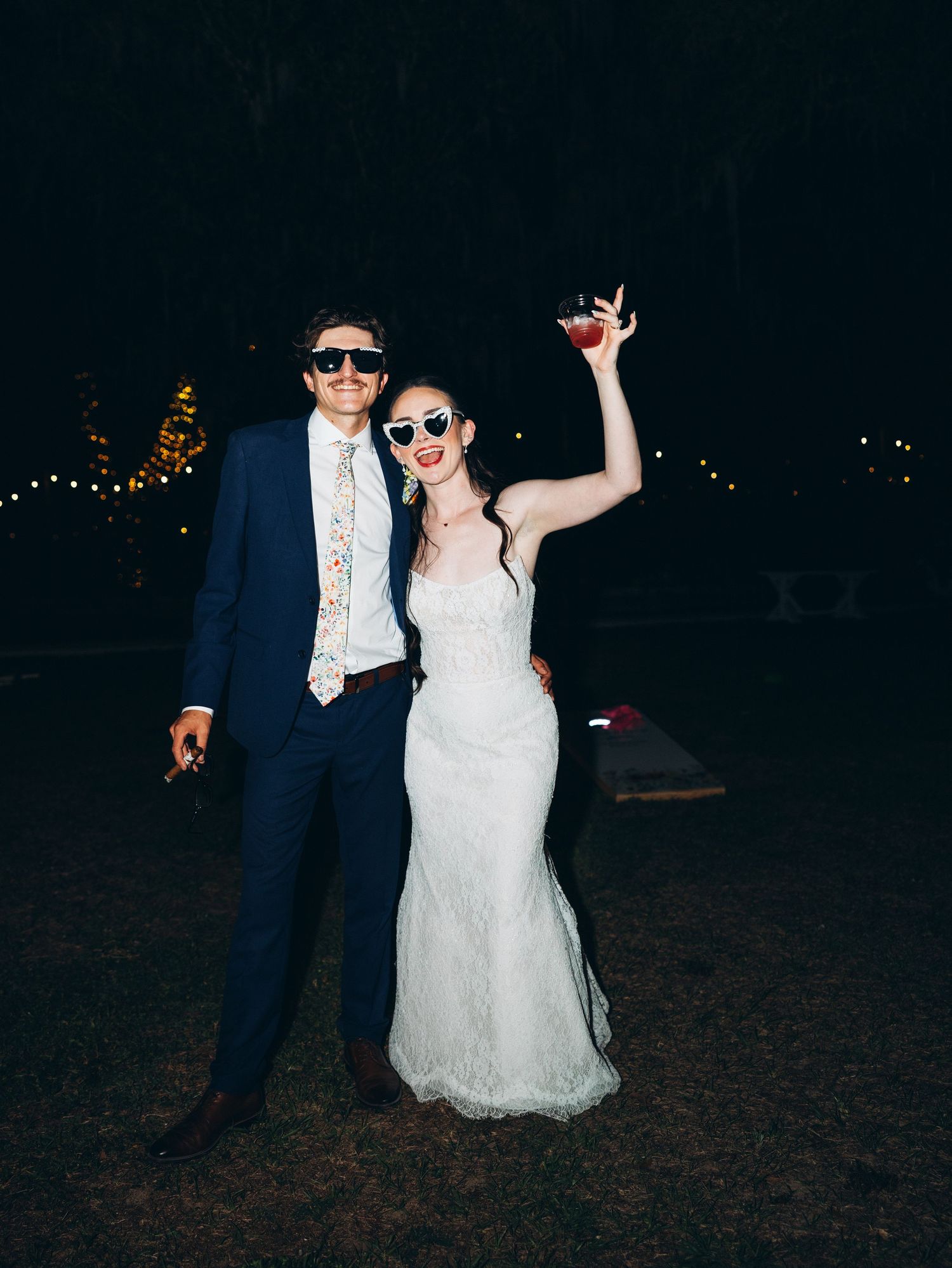 Bride in white lace gown and groom in navy suit wearing sunglasses, celebrating outdoors at night with drinks raised.