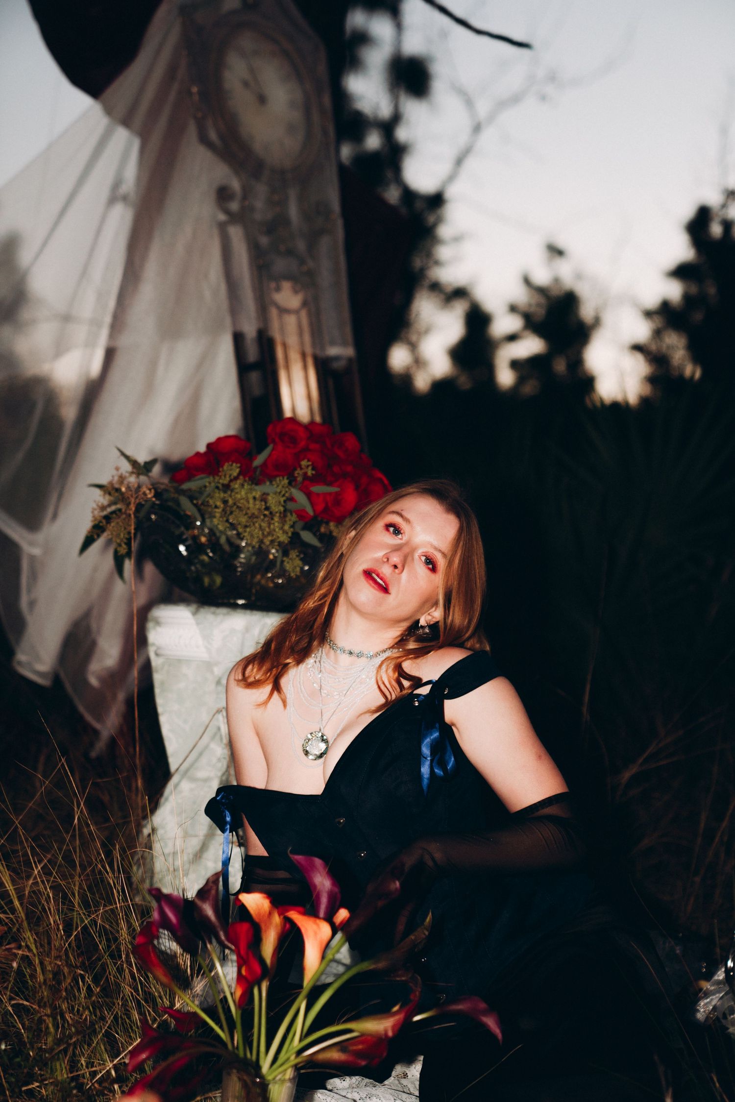 Woman in black gown with necklace poses among flowers and clock at dusk in gothic outdoor setting.