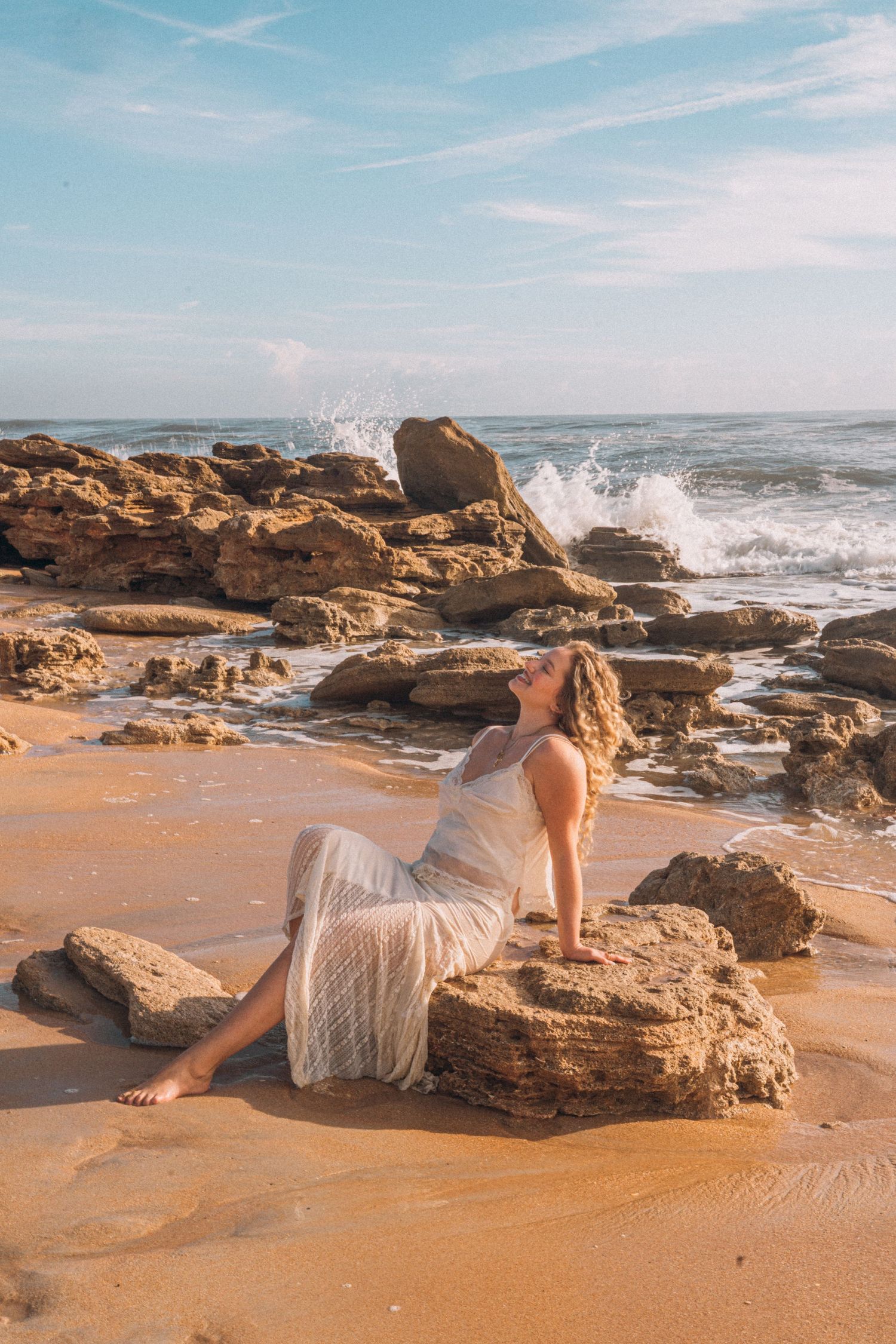 Woman in white dress sitting on rocky beach with ocean waves crashing behind her on a sunny day.