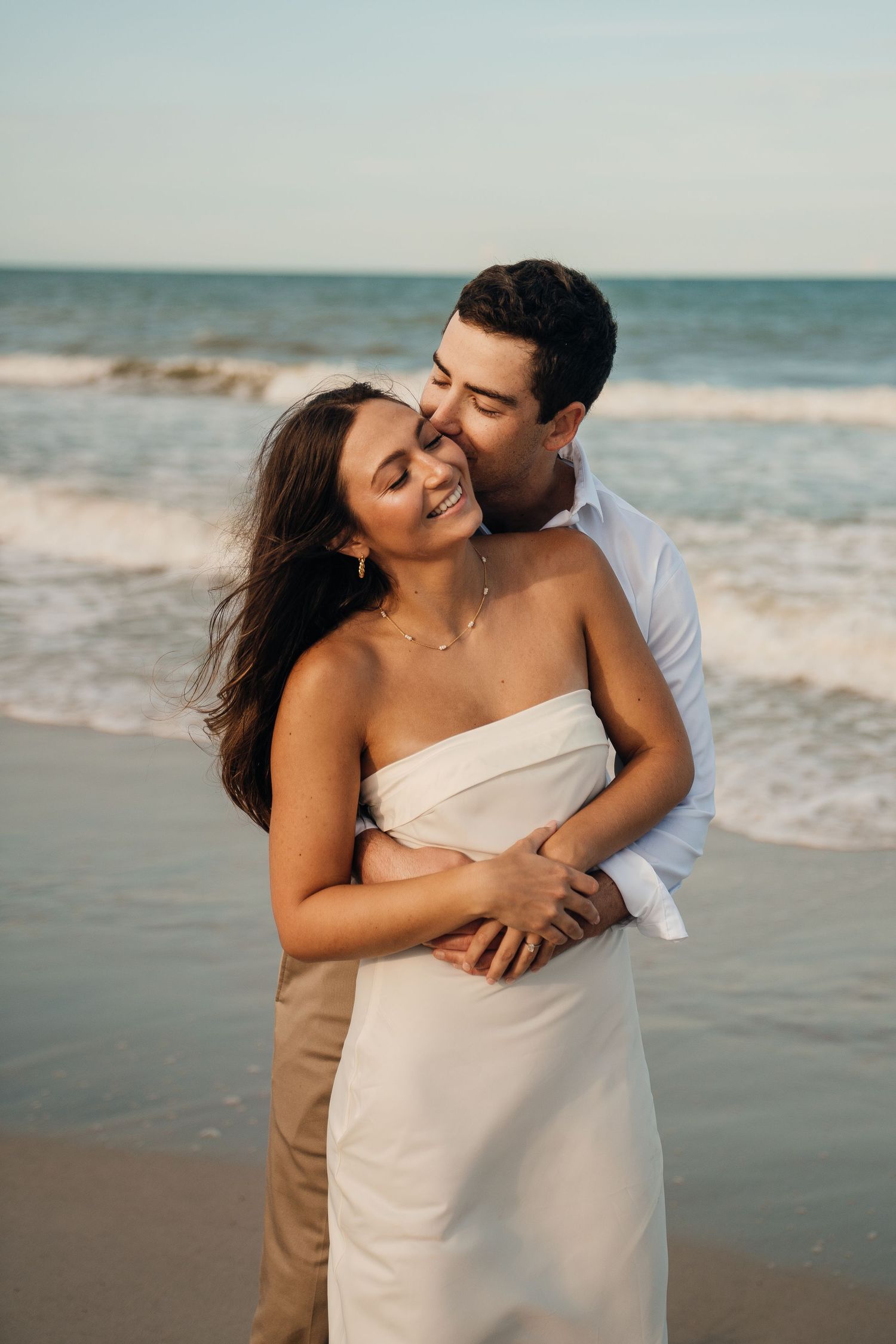 A tender embrace between a couple on the beach with crashing waves in the background.