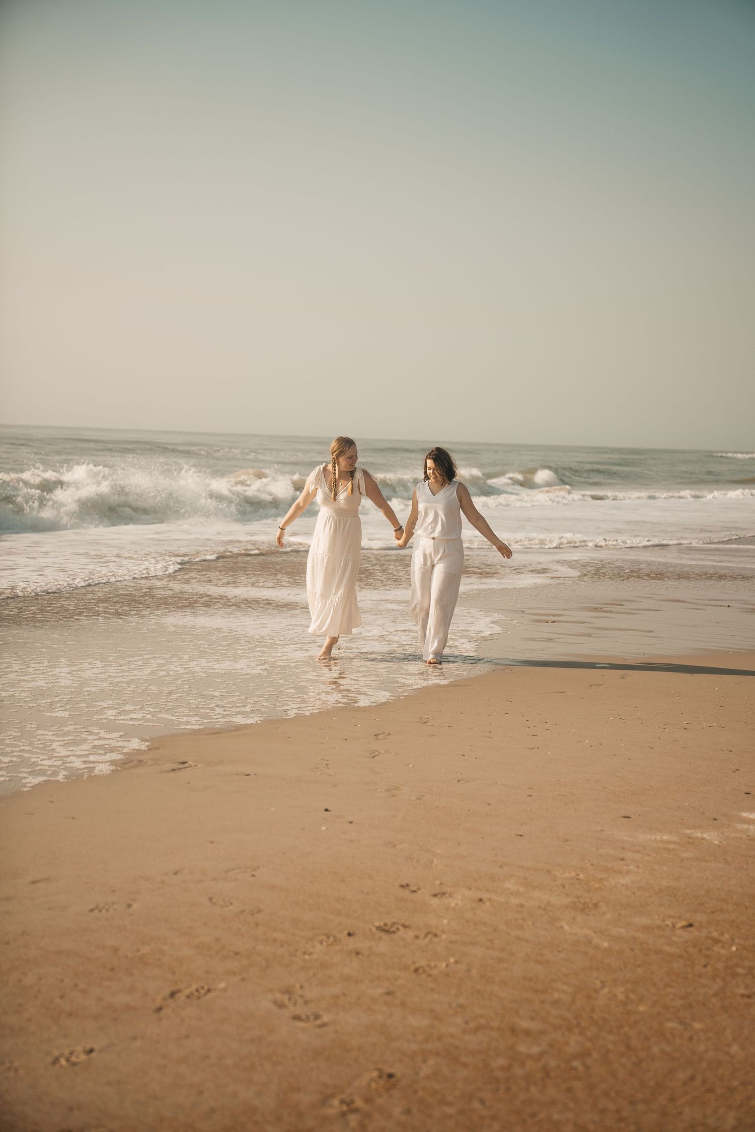Two figures in white dresses run playfully along a sandy beach with ocean waves in the background.