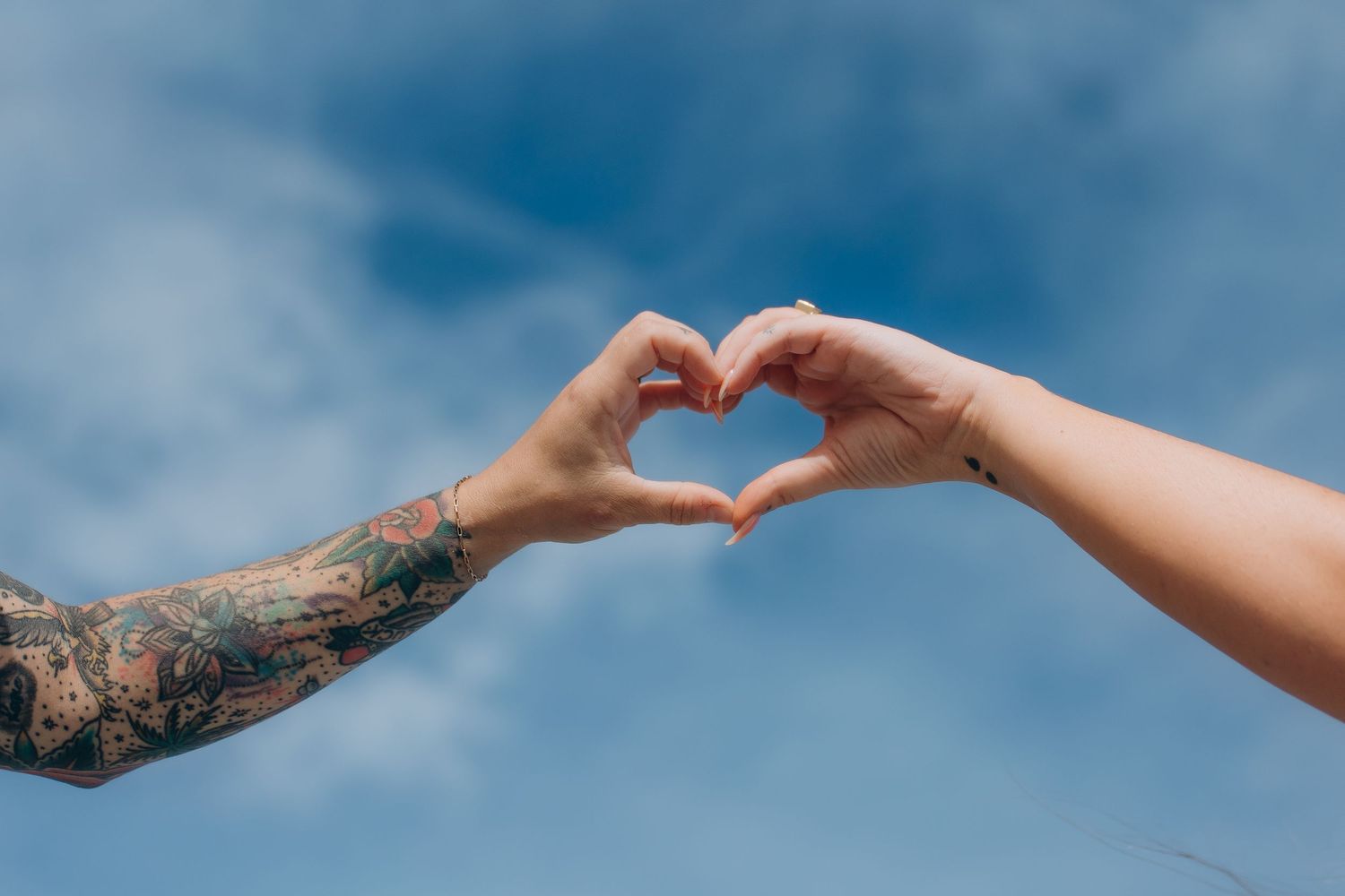 Hands forming a heart shape against a bright blue sky with clouds.