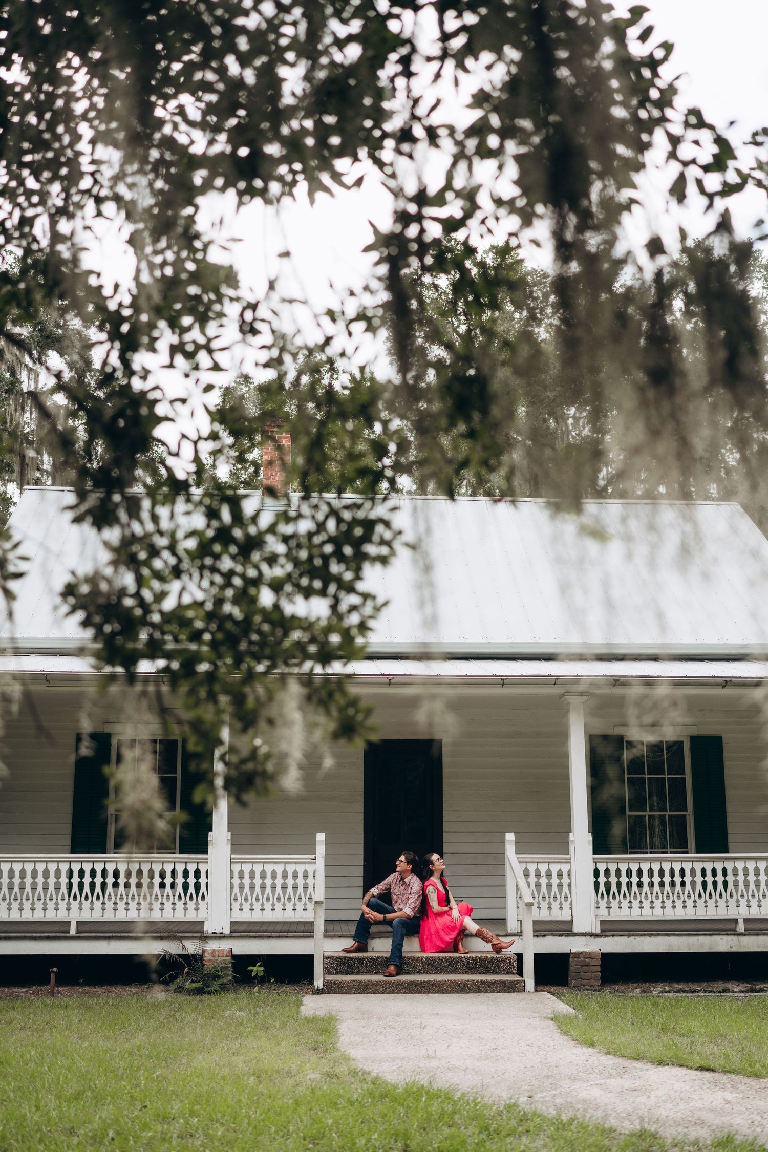 White plantation-style house with a wraparound porch and black shutters under moss-draped oak trees.