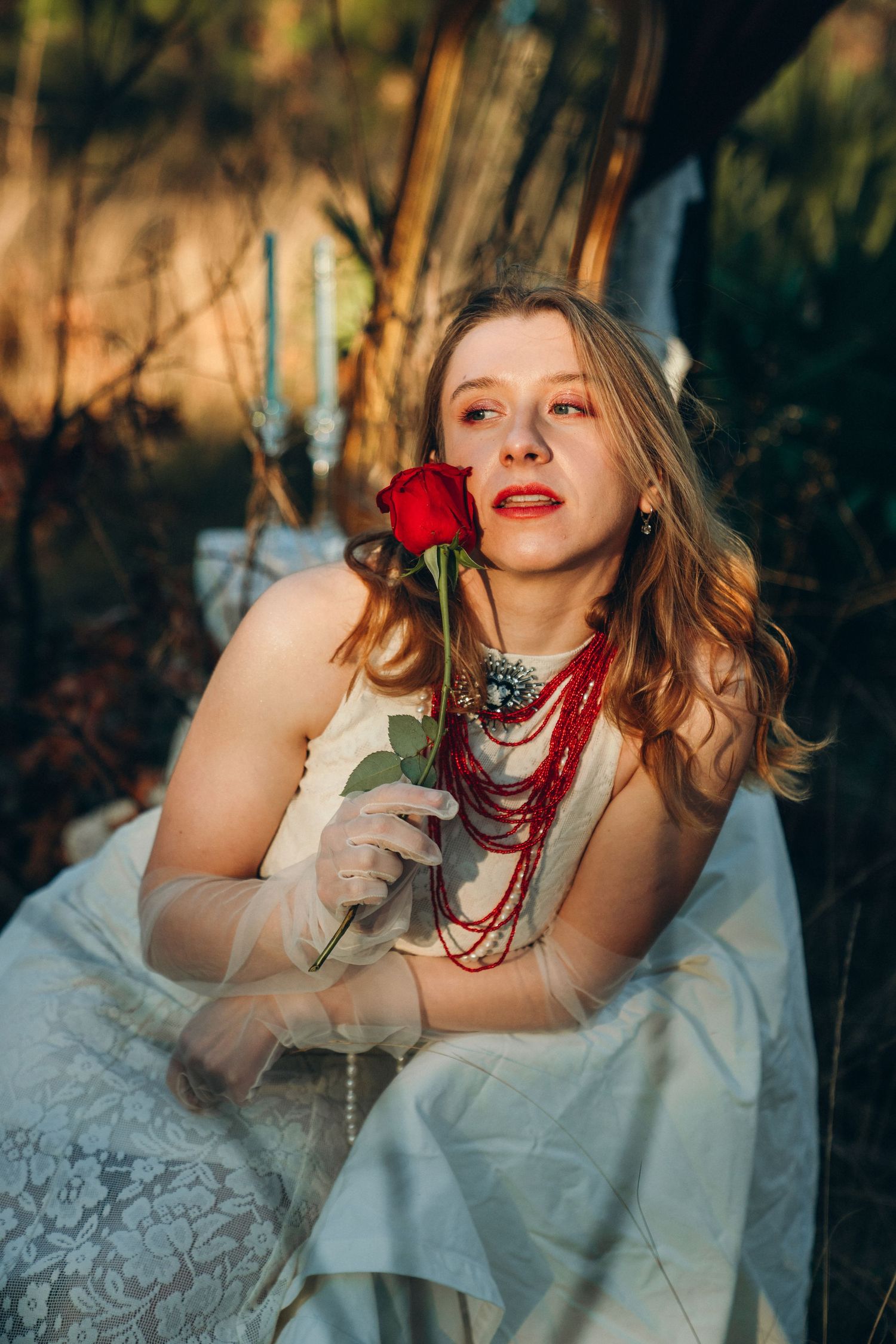 Young woman in white lace dress holding red rose, wearing red beaded necklace and gloves, seated outdoors.