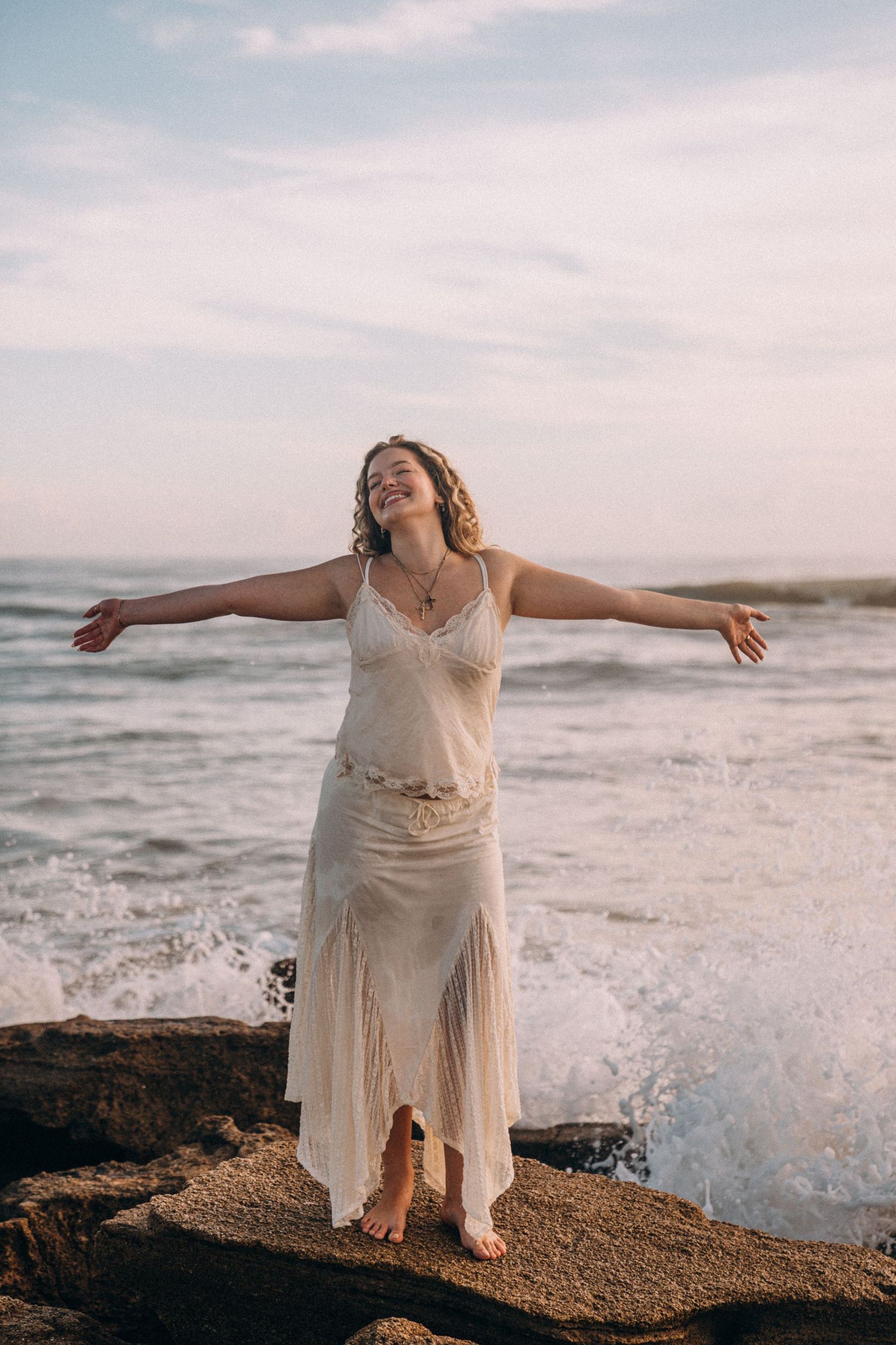 Woman in white dress standing on rocks with arms outstretched, smiling at ocean waves at sunset.