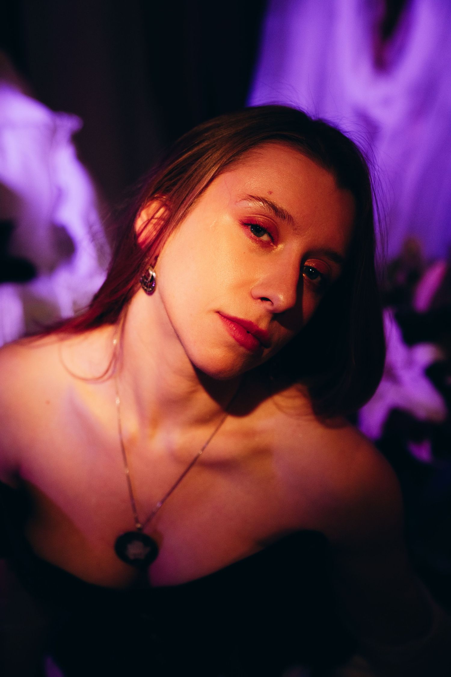 Young woman with red lips and a pendant necklace poses under moody purple and red lighting.