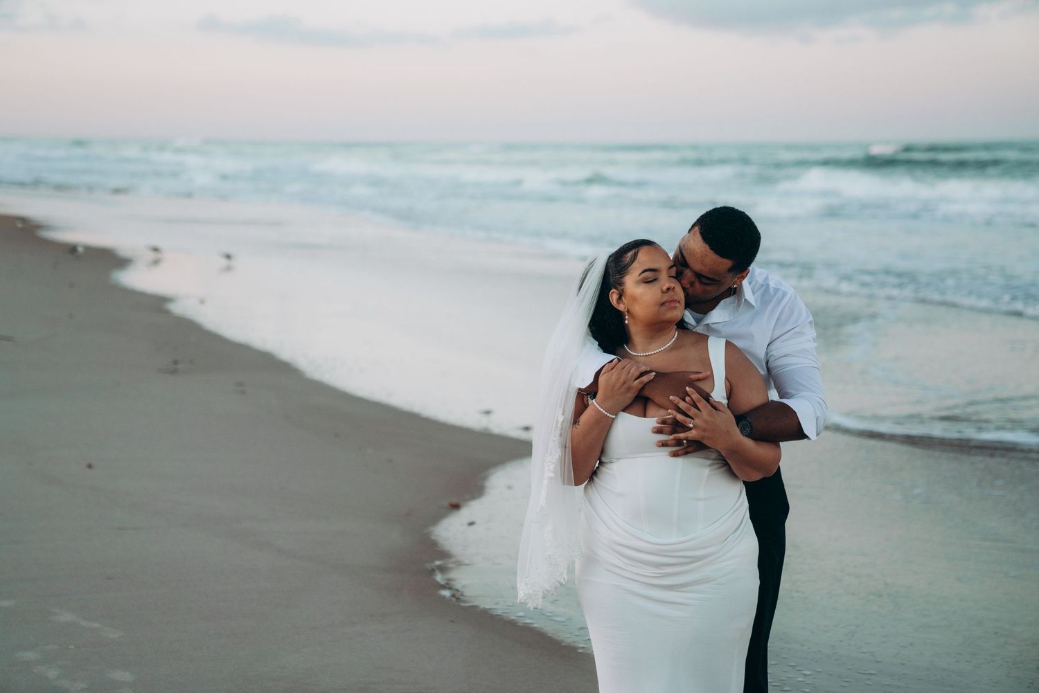 Couple in wedding attire embracing on a beach at sunset, bride in white gown with veil, groom in black suit.